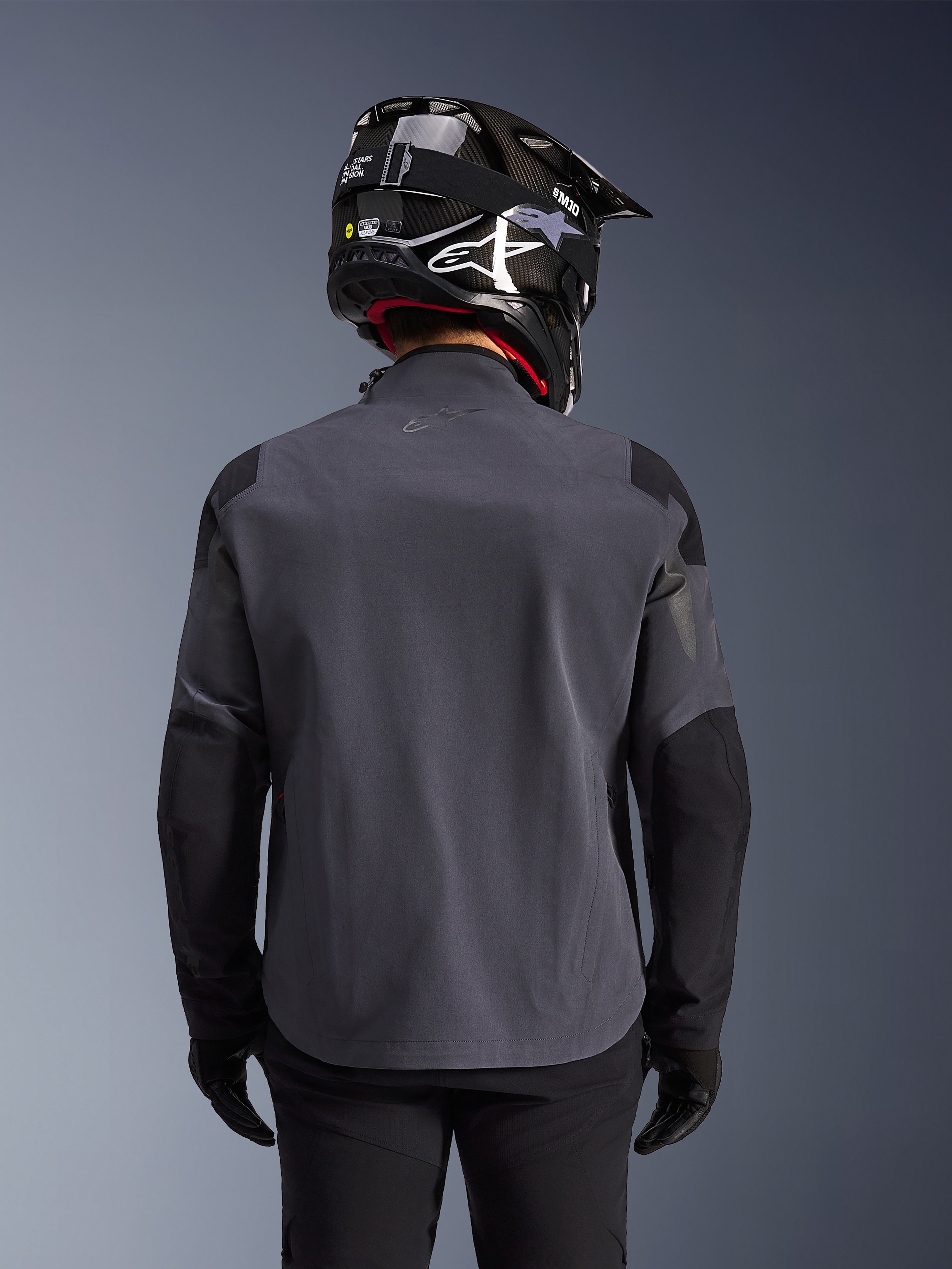 Maxdura Drystar®XF Laminierte Gepanzerte Jacke, Adventure-Motorradjacke, Dunkelgrau Schwarz, Rückansicht mit technischer Textilkonstruktion und verstärkten Panelen, getragen mit schwarzem Carbon-Helm, schwarzer Fahrhose und Handschuhen