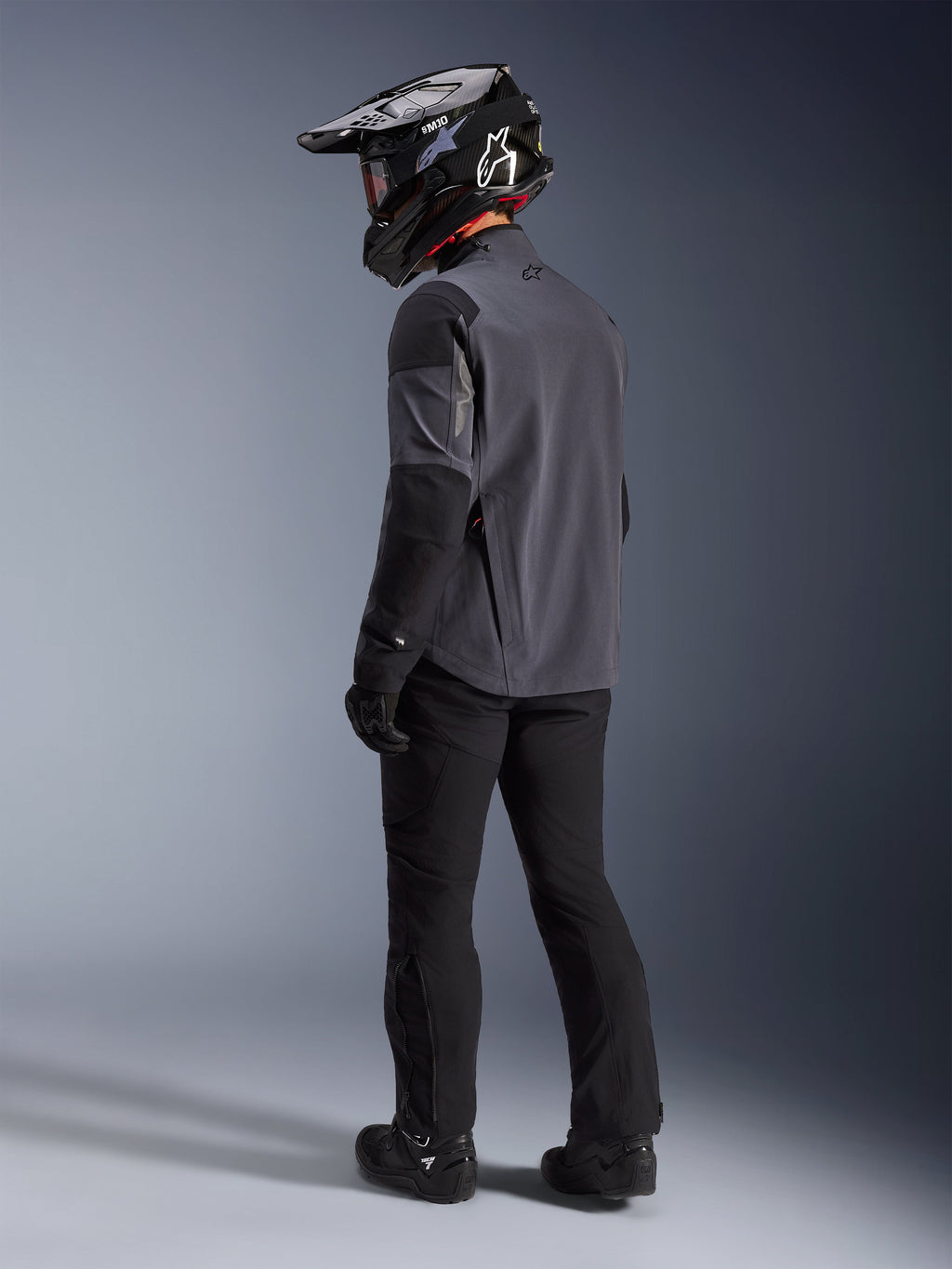 Adventure-Fahrer in der Maxdura Drystar®XF Laminierten Gepanzerten Schale in Deep Gray Black, Rückansicht. Komplette Ausrüstung inklusive schwarzer Motorradhose, einem schwarzen Motocross-Helm mit Alpinestars-Branding und schwarzen Offroad-Stiefeln. Technische Allwetter-Fahrausrüstung für Dual-Sport-Performance.