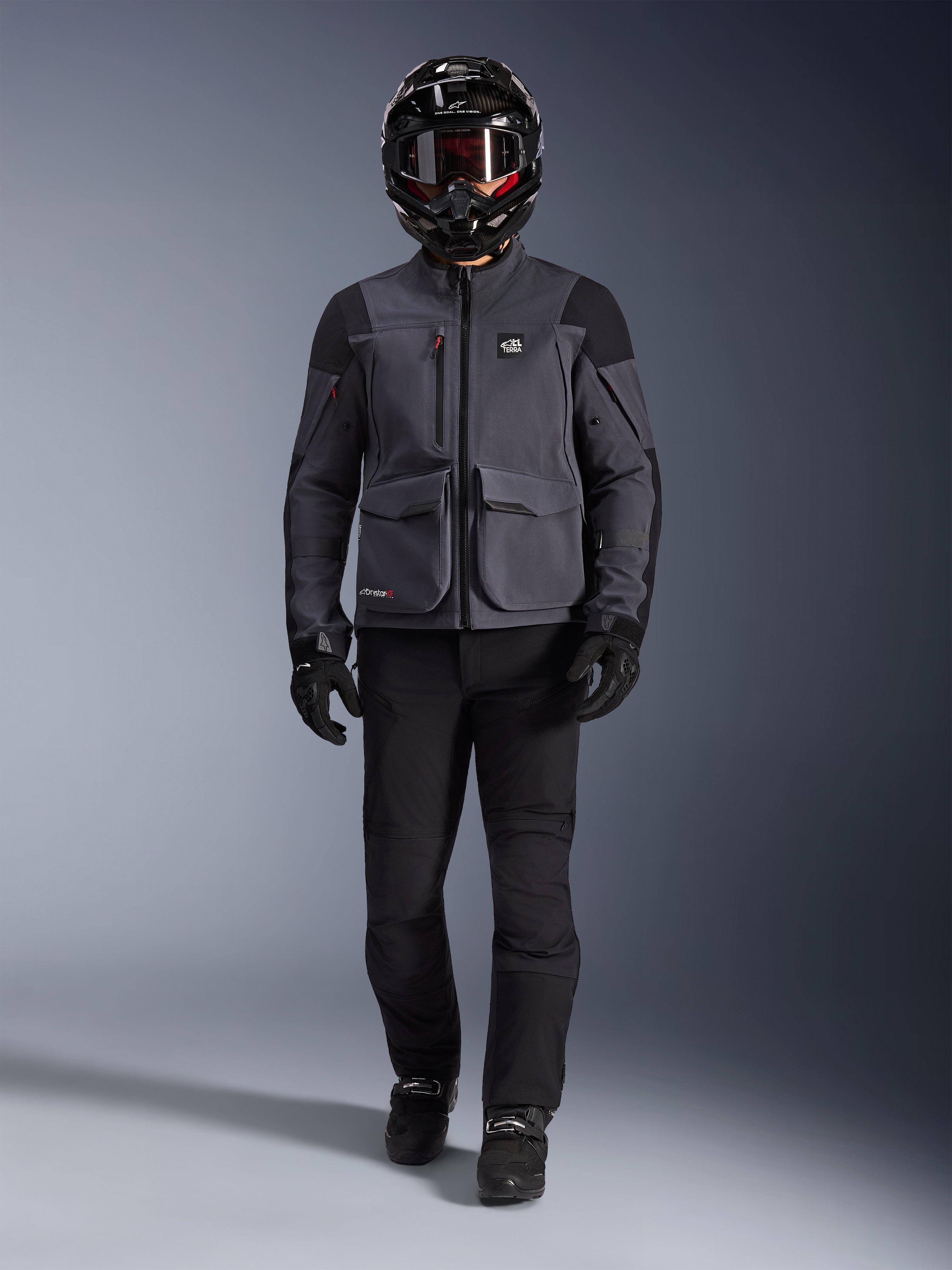 Alpinestars Maxdura Drystar®XF Laminierte Gepanzerte Jacke, Adventure-Motorradjacke in Deep Gray und Schwarz, getragen mit passender schwarzer Motorradhose, einem schwarzen Integralhelm, Handschuhen und Stiefeln, mit markanten Cargotaschen und ALL TERRA Branding für einen technischen Offroad-Touren-Look