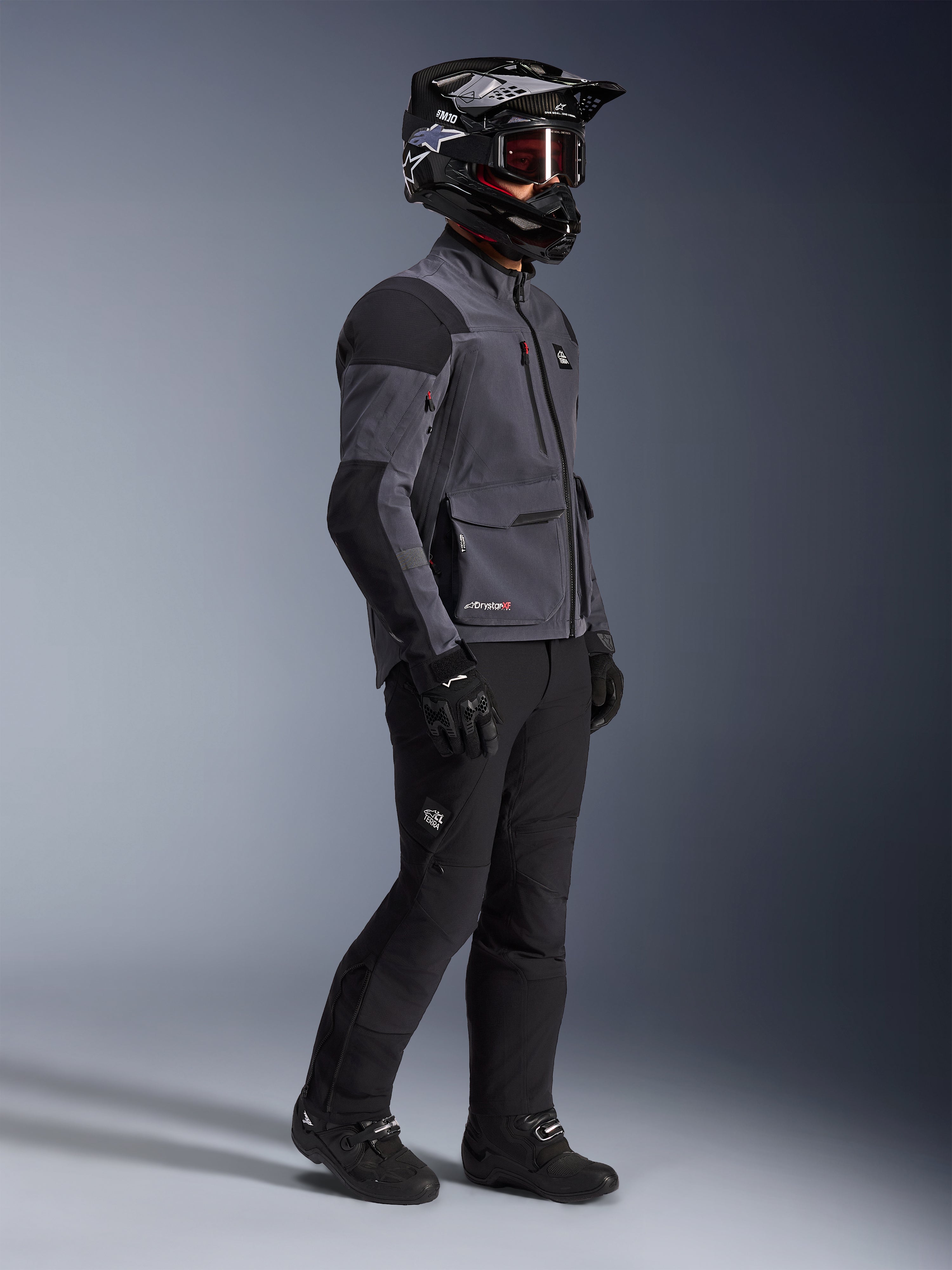 Motorradfahrer mit Alpinestars Maxdura Drystar®XF Laminated Armored Shell Jacke in Deep Gray und Schwarz, kombiniert mit schwarzer Adventure-Fahrhose, schwarzem Helm mit Brille, schwarzen Handschuhen und Stiefeln, Ganzkörper-Studioaufnahme zur Präsentation der technischen Dual-Sport-Ausrüstung.