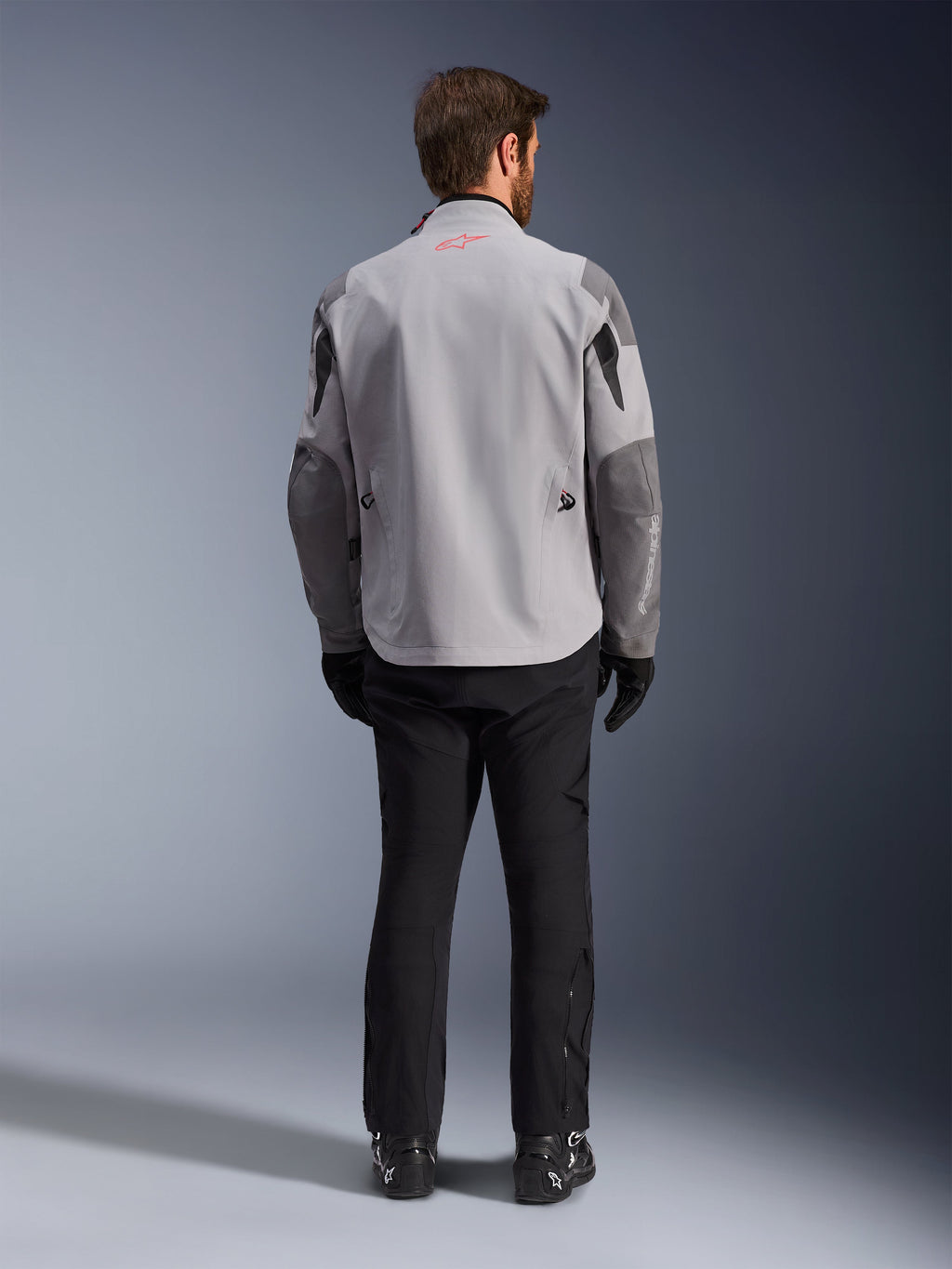 Alpinestars Maxdura Drystar®XF Laminierte Gepanzerte Jacke, Motorradjacke in Mittelgrau und Dunkelgrau, Rückansicht, mit hellgrauem Hauptteil und dunkelgrauen Einsätzen an Schultern und Ärmeln, kleinem roten Alpinestars-Sternlogo am hinteren Kragen und weißem Branding am unteren linken Ärmel, kombiniert mit schwarzer Motorradhose und Stiefeln.