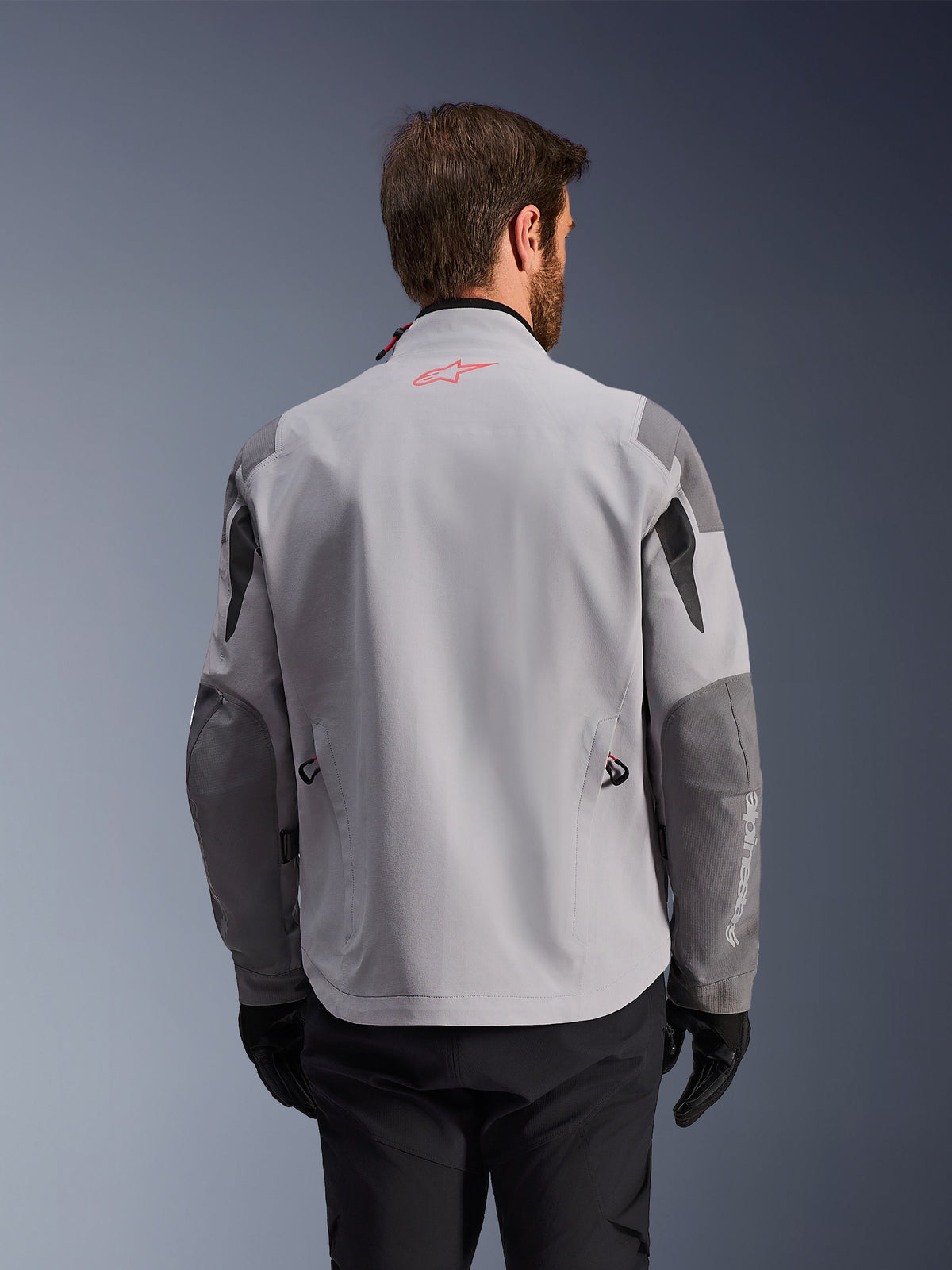 Alpinestars Maxdura Drystar®XF Laminierte Gepanzerte Jacke, Rückansicht einer Adventure-Motorradjacke, Mittelgrau und Dunkelgrau, mit einem roten Stern-Logo am Kragen und weißem vertikalen Branding am Ärmel, kombiniert mit schwarzen Motorradhandschuhen und -hosen