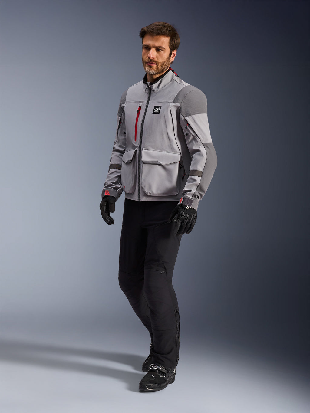 Alpinestars Maxdura Drystar®XF Laminierte Gepanzerte Jacke, Adventure-Motorradjacke, Mittelgrau und Dunkelgrau, getragen von einem Model mit schwarzen Motorradhosen und Handschuhen, mit roten Reißverschluss-Akzenten und Cargotaschen, professionelle technische Allwetter-Ausrüstung für Adventure-Touren