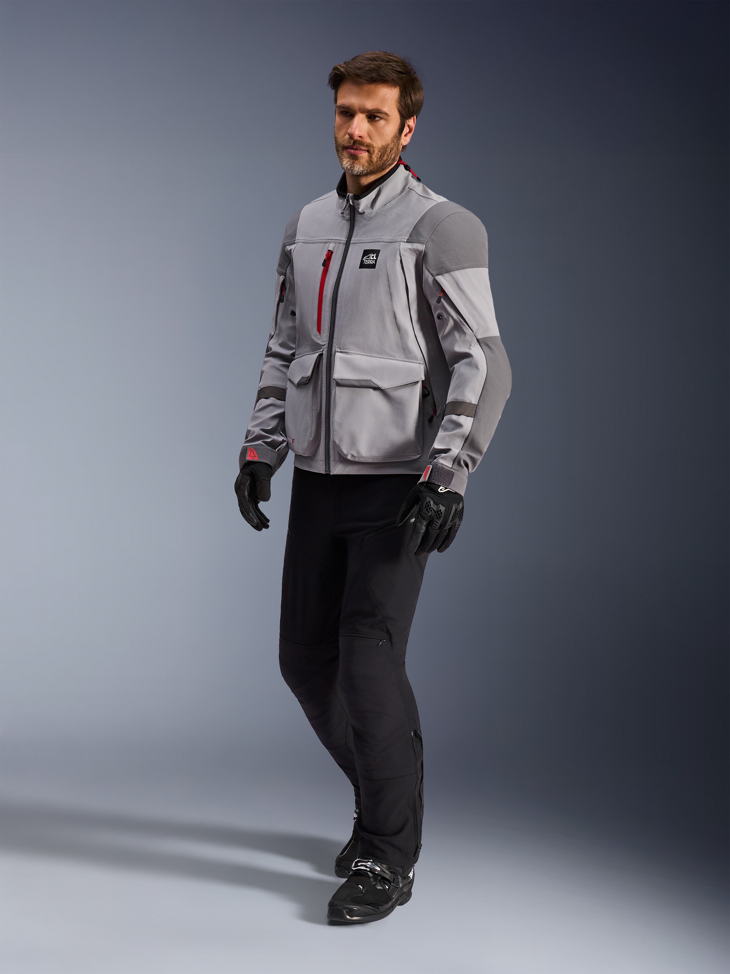 Alpinestars Maxdura Drystar®XF Laminierte Gepanzerte Jacke, Adventure-Motorradjacke, Mittelgrau und Dunkelgrau, getragen von einem Model mit schwarzen Motorradhosen und Handschuhen, mit roten Reißverschluss-Akzenten und Cargotaschen, professionelle technische Allwetter-Ausrüstung für Adventure-Touren