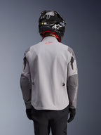 Alpinestars Maxdura Drystar®XF Laminierte Gepanzerte Jacke, Adventure-Motorradjacke, Mittelgrau und Dunkelgrau, Rückansicht mit hellgrauem Hauptteil und dunkelgrauen verstärkten Einsätzen an Schultern und Ellbogen, rotes Alpinestars-Logo am oberen Rücken, getragen mit schwarzem Helm und schwarzen Handschuhen.