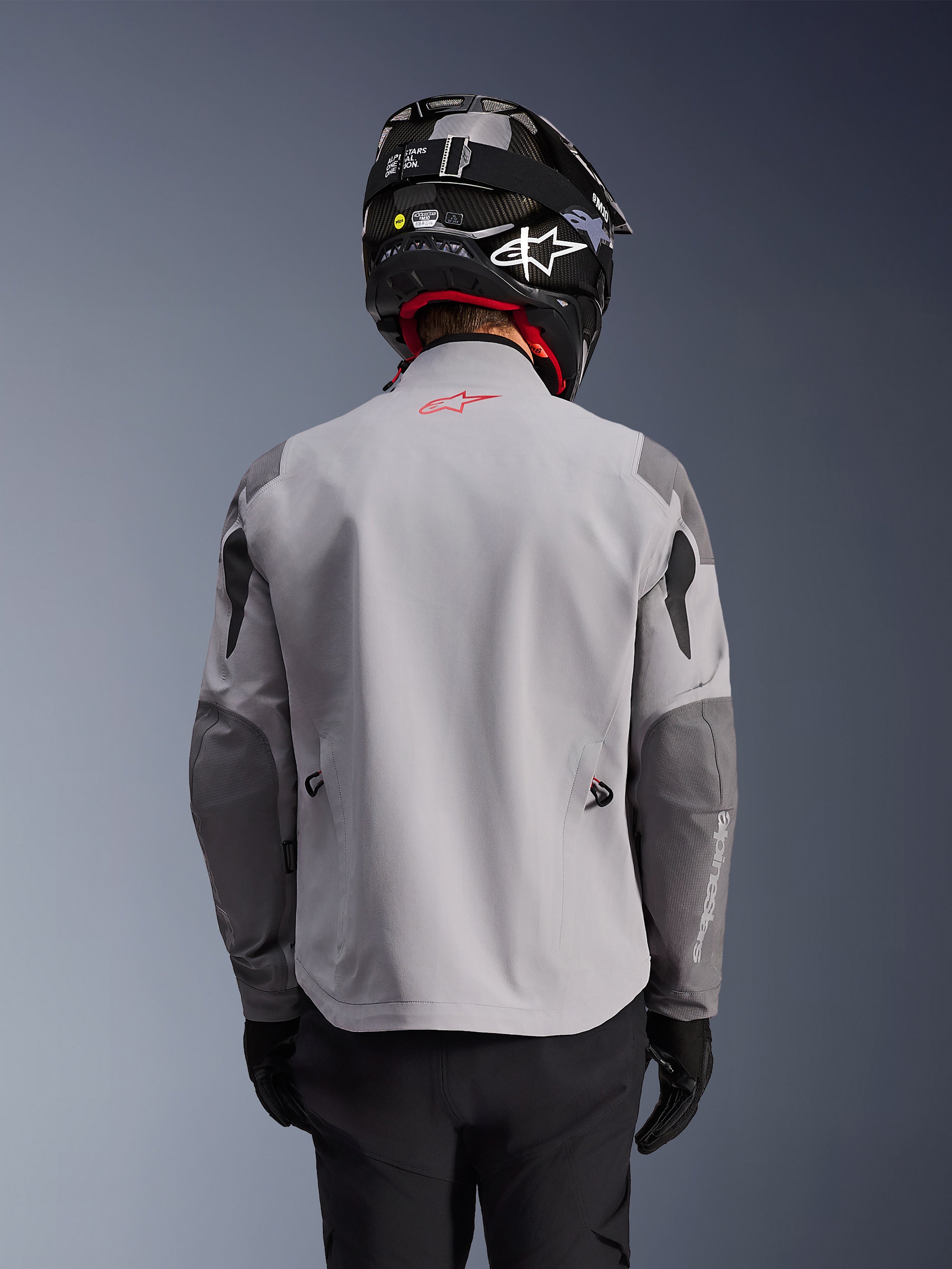 Alpinestars Maxdura Drystar®XF Laminierte Gepanzerte Jacke, Adventure-Motorradjacke, Mittelgrau und Dunkelgrau, Rückansicht mit hellgrauem Hauptteil und dunkelgrauen verstärkten Einsätzen an Schultern und Ellbogen, rotes Alpinestars-Logo am oberen Rücken, getragen mit schwarzem Helm und schwarzen Handschuhen.