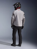 Rückansicht eines Motorradfahrers in der Alpinestars Maxdura Drystar®XF Laminated Armored Shell Jacke in Mittelgrau und Dunkelgrau, mit einem roten Stern-Logo am Kragen, kombiniert mit einer schwarzen Adventure-Hose, einem schwarzen Helm mit Carbon-Muster und schwarzen Stiefeln.