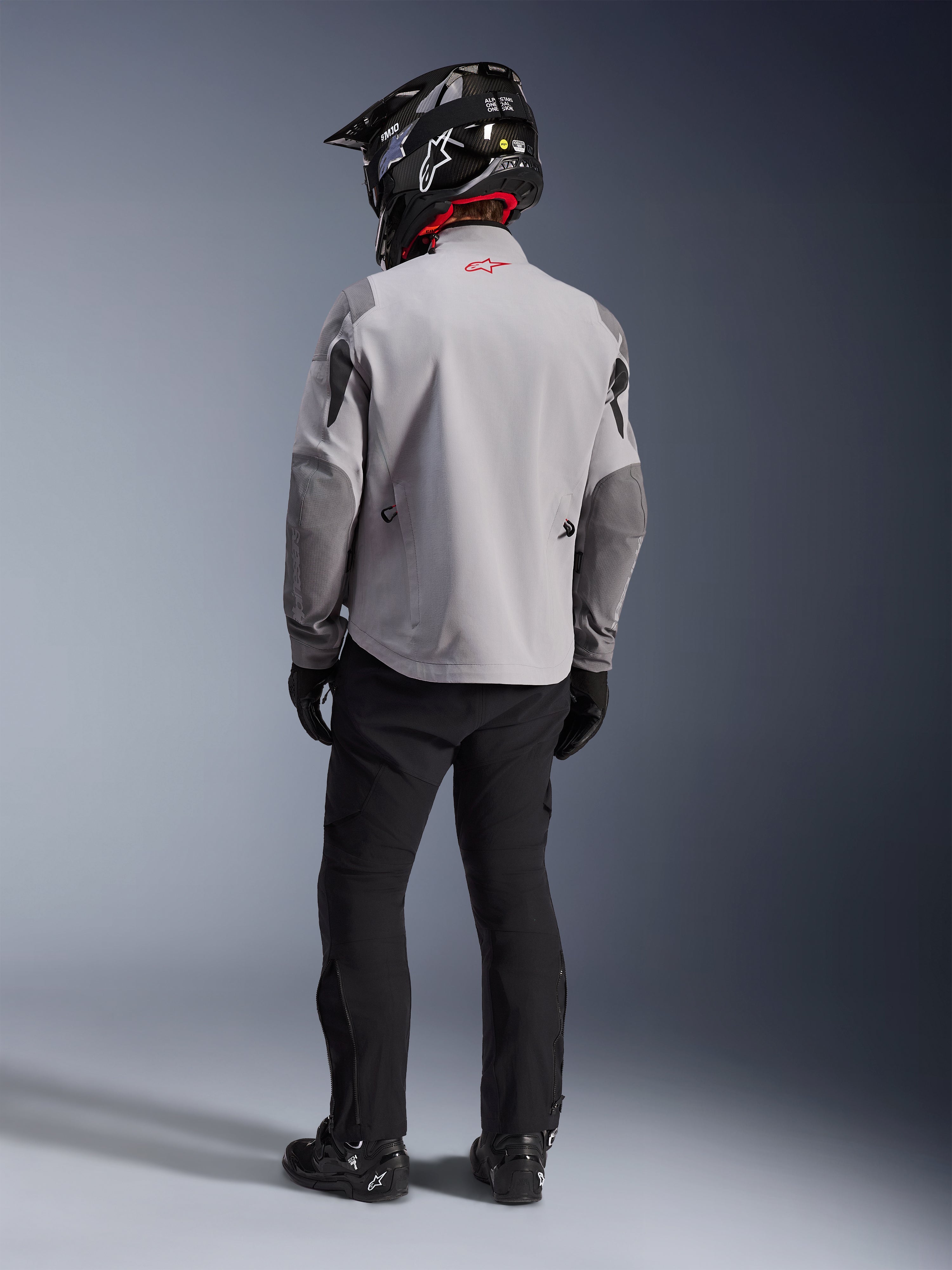 Rückansicht eines Motorradfahrers in der Alpinestars Maxdura Drystar®XF Laminated Armored Shell Jacke in Mittelgrau und Dunkelgrau, mit einem roten Stern-Logo am Kragen, kombiniert mit einer schwarzen Adventure-Hose, einem schwarzen Helm mit Carbon-Muster und schwarzen Stiefeln.