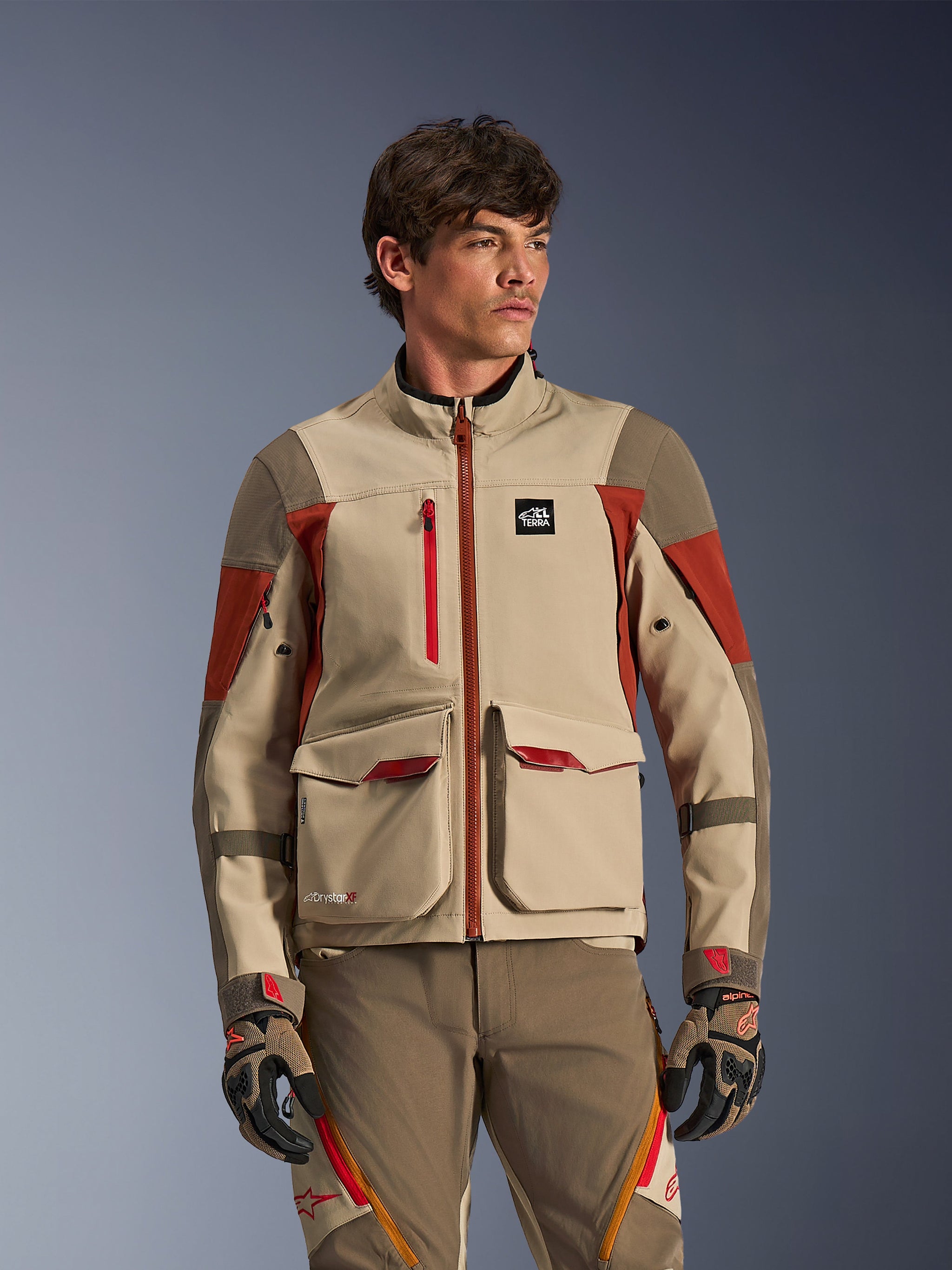 Alpinestars Maxdura Drystar®XF Laminated Armored Shell, Adventure-Motorradjacke in Aluminum Walnut Rust Brown, getragen von einem männlichen Model, mit großen vorderen Cargo-Taschen und technischen roten Akzenten, kombiniert mit passender Adventure-Fahrhose und Handschuhen