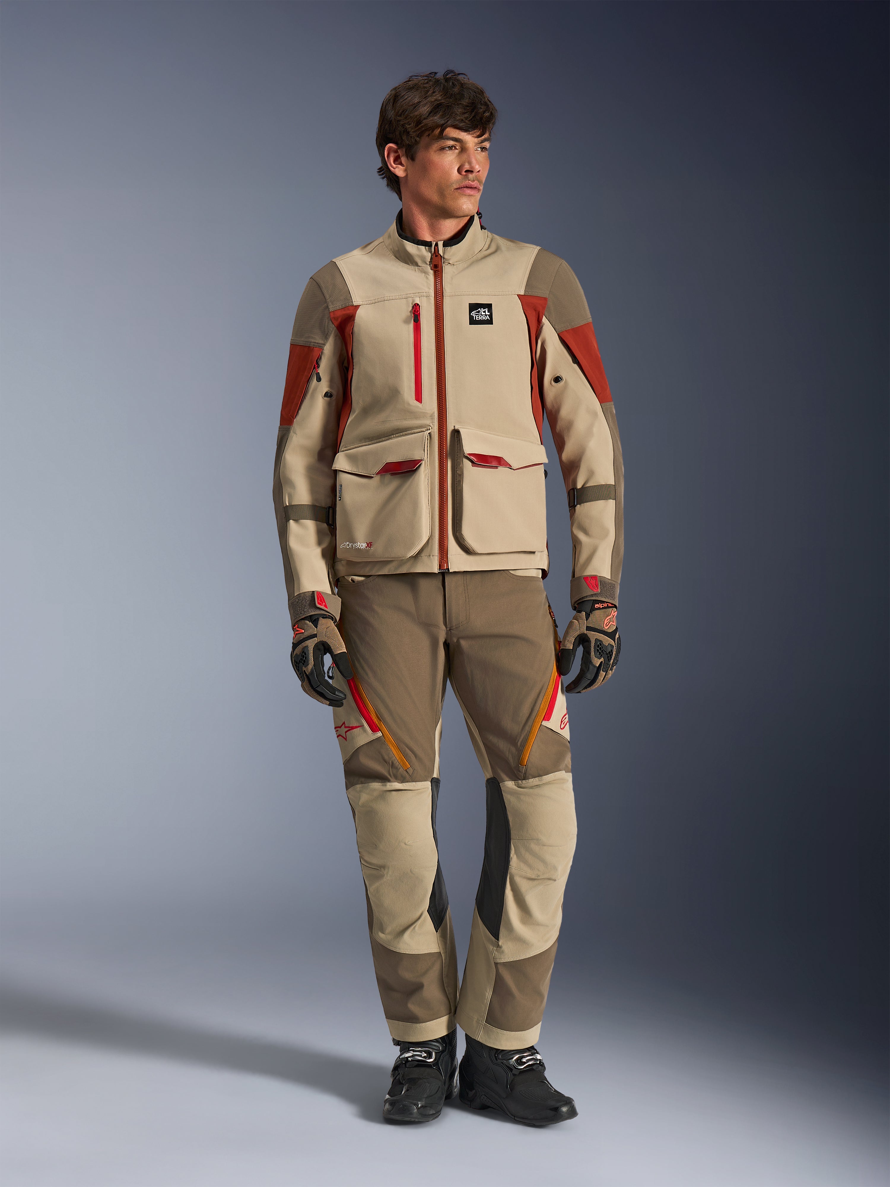 Maxdura Drystar®XF Laminated Armored Shell, Adventure-Motorradjacke in Aluminum Walnut Rust Brown, getragen von einem männlichen Model mit passender Adventure-Hose, Handschuhen und Stiefeln, Ganzkörper-Studioaufnahme einer professionellen Tourenausrüstung.