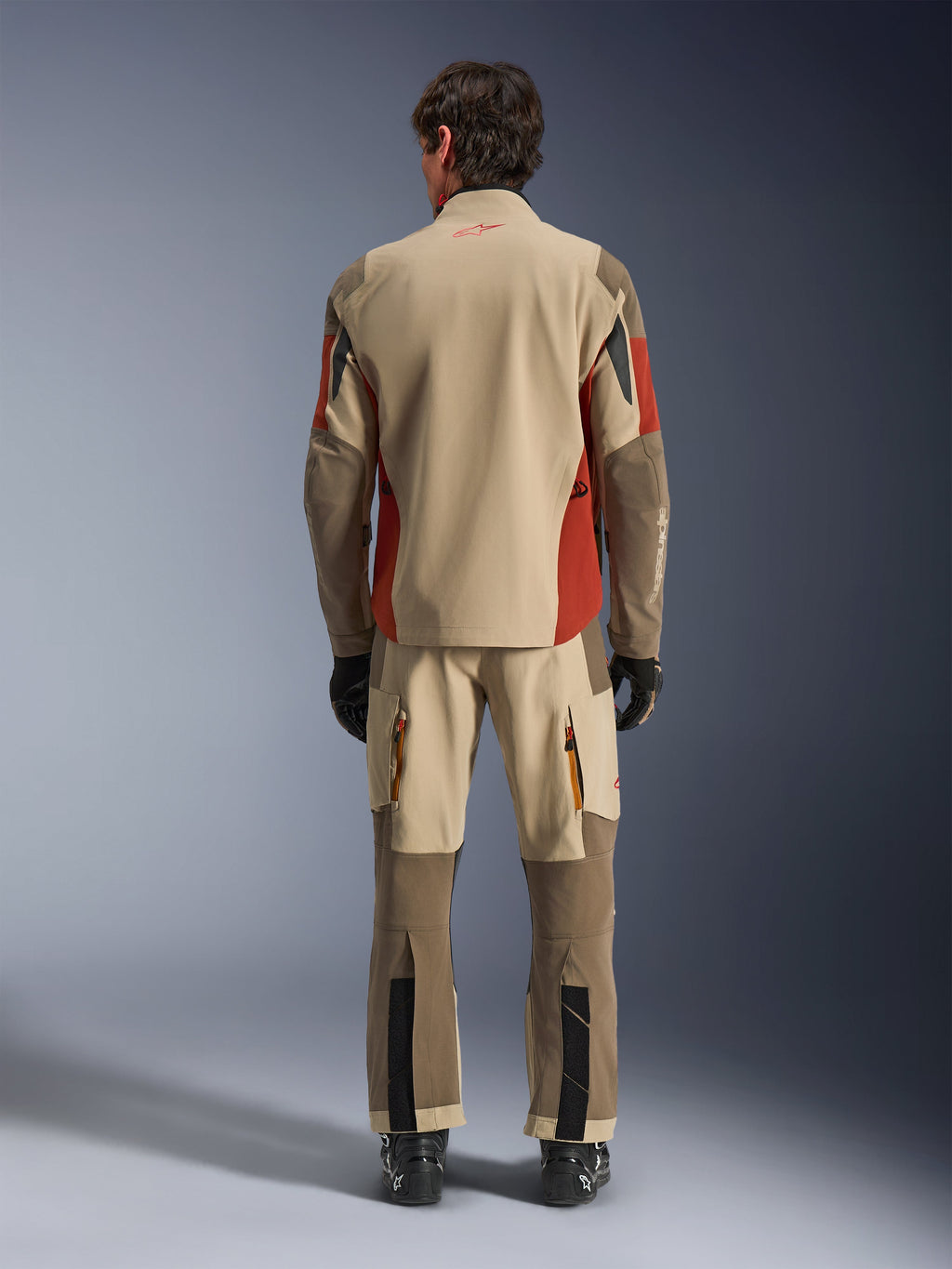 Rückansicht eines Fahrers in der Alpinestars Maxdura Drystar®XF Laminated Armored Shell und passender Adventure-Hose in Aluminum Walnut Rust Brown, beigefarbenes und braunes Textil mit orangefarbenen Akzenten, kombiniert mit schwarzen Motorradstiefeln und Handschuhen, professionelle Adventure-Tourenausrüstung.