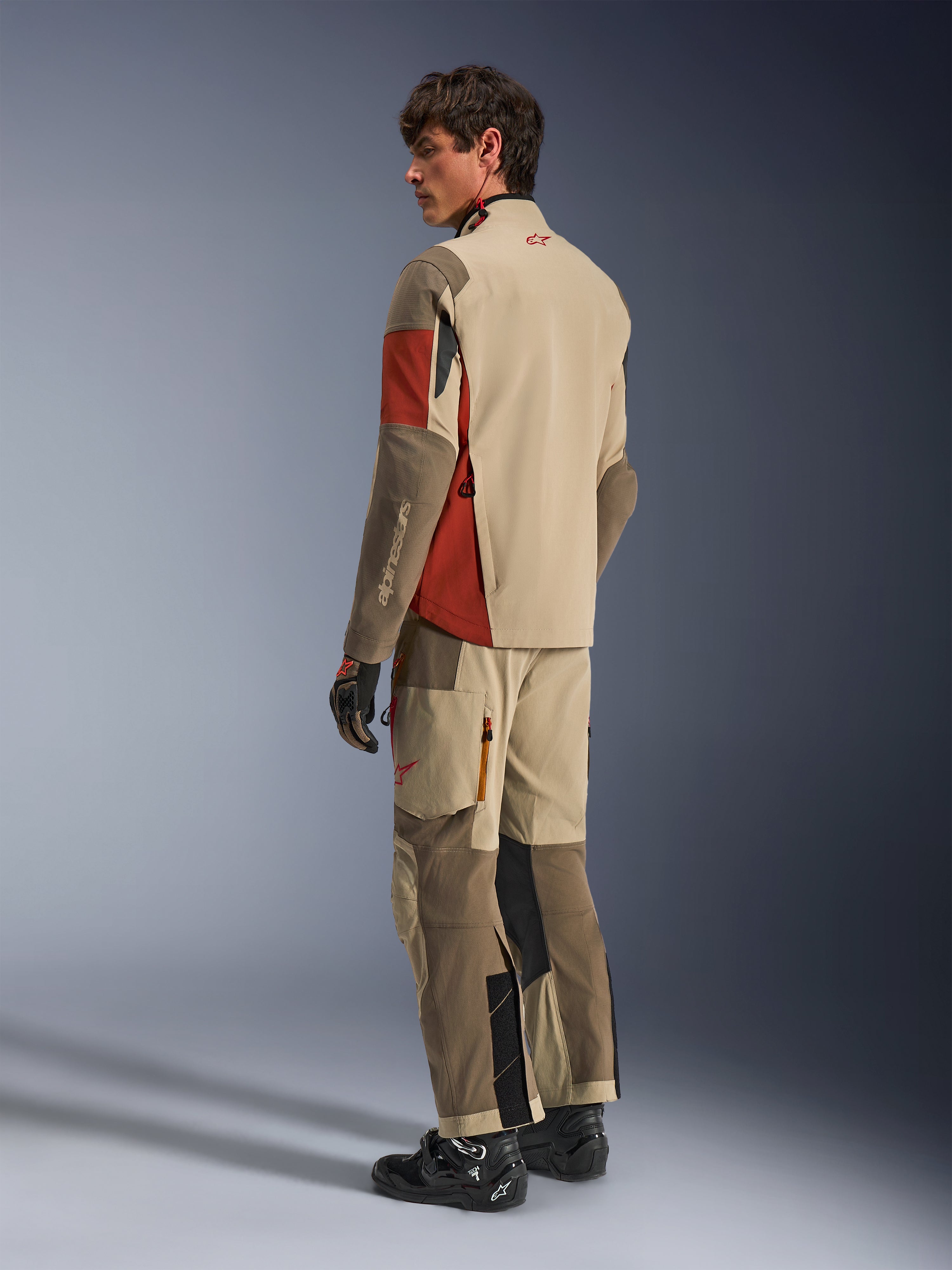 Männliches Model in der Alpinestars Maxdura Drystar®XF laminierten gepanzerten Schale in Aluminum Walnut Rust Brown, dazu passende Adventure-Tourenhose, schwarze Handschuhe und schwarze Stiefel; Rückansicht, die die technische Textilkonstruktion und die verstärkten Zonen zeigt.