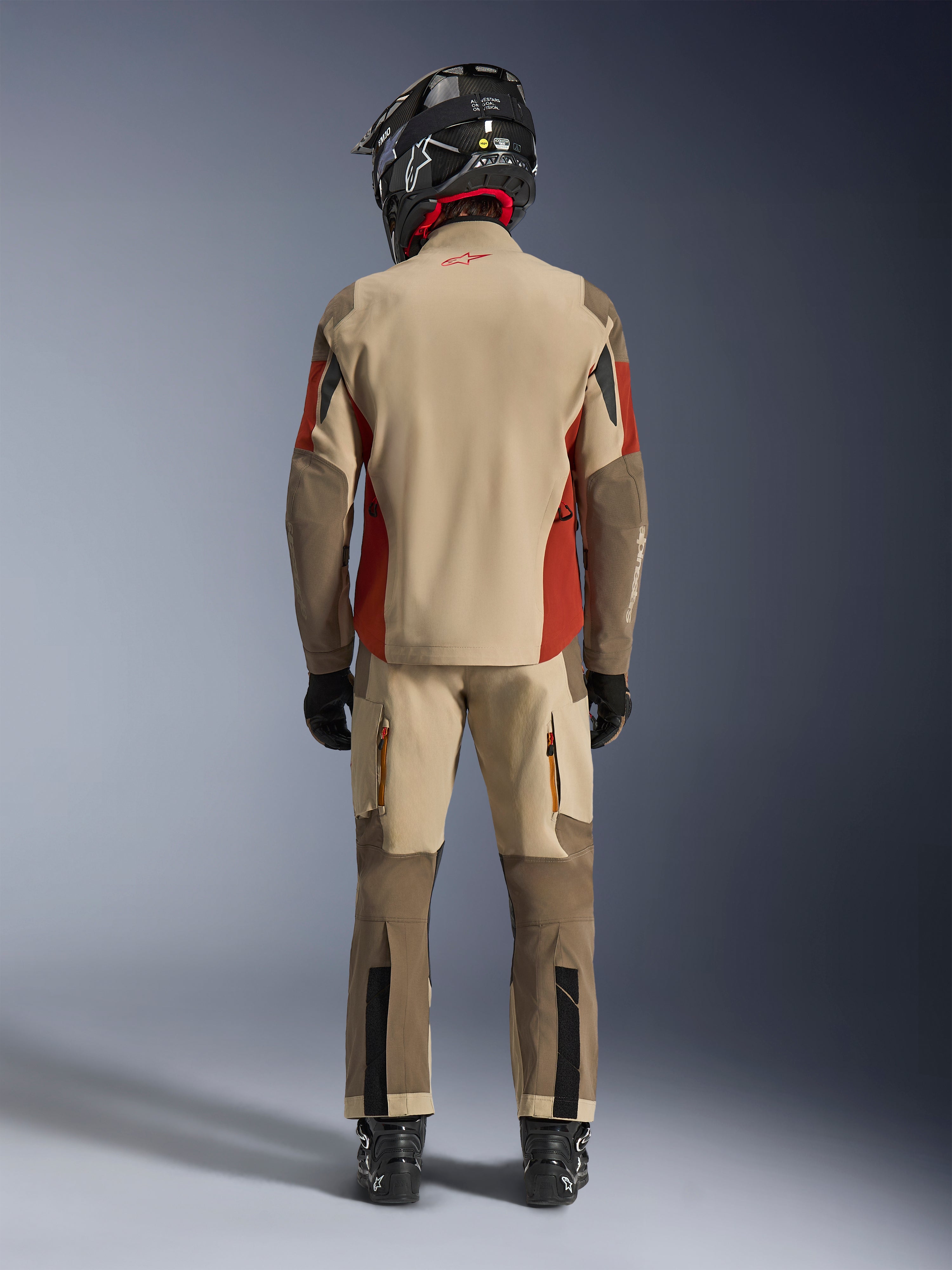 Alpinestars Maxdura Drystar®XF Laminated Armored Shell, Adventure-Motorradjacke und passende Hose, Farbgebung Aluminum Walnut und Rust Brown, Rückansicht eines Fahrers mit schwarzem Helm und Stiefeln, professionelle Adventure-Touring-Ausrüstung