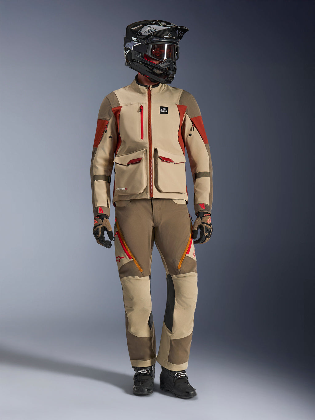 Ganzkörper-Studioaufnahme eines Fahrers in der Alpinestars Maxdura Drystar®XF Laminated Armored Shell Jacke in Aluminum Walnut Rust Brown, kombiniert mit einer passenden Adventure-Hose, einem schwarzen Offroad-Helm mit rot getönter Brille und technischen Handschuhen; zeigt ein komplettes Adventure-Touring-Outfit.