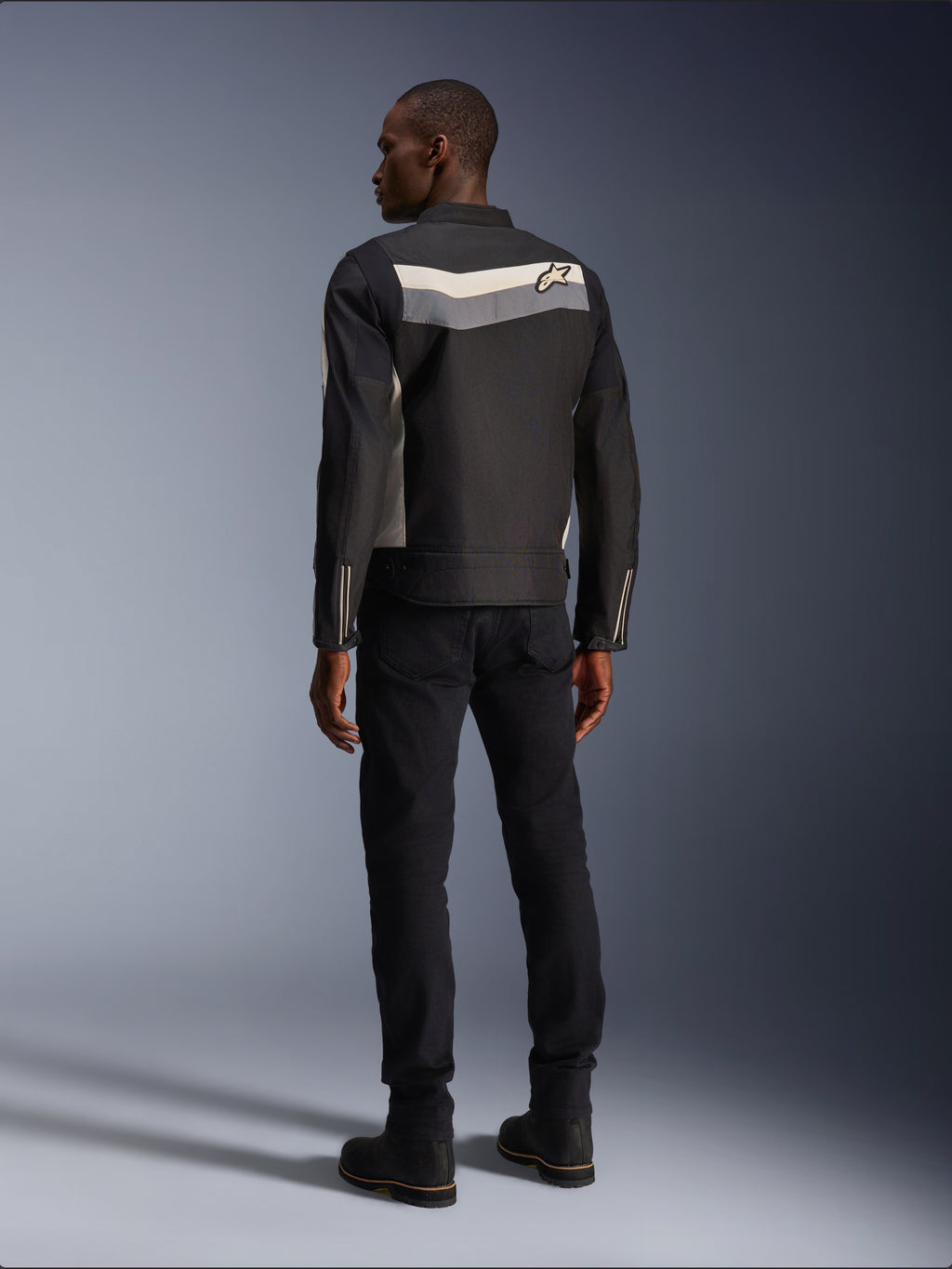 Male model from behind wearing the Alpinestars T-Dyno WR Jacket in Black Dark Gray Ecru, textile motorcycle jacket with a horizontal stripe and star logo on the back, paired with black riding pants and boots, urban motorcycle gear aesthetic.