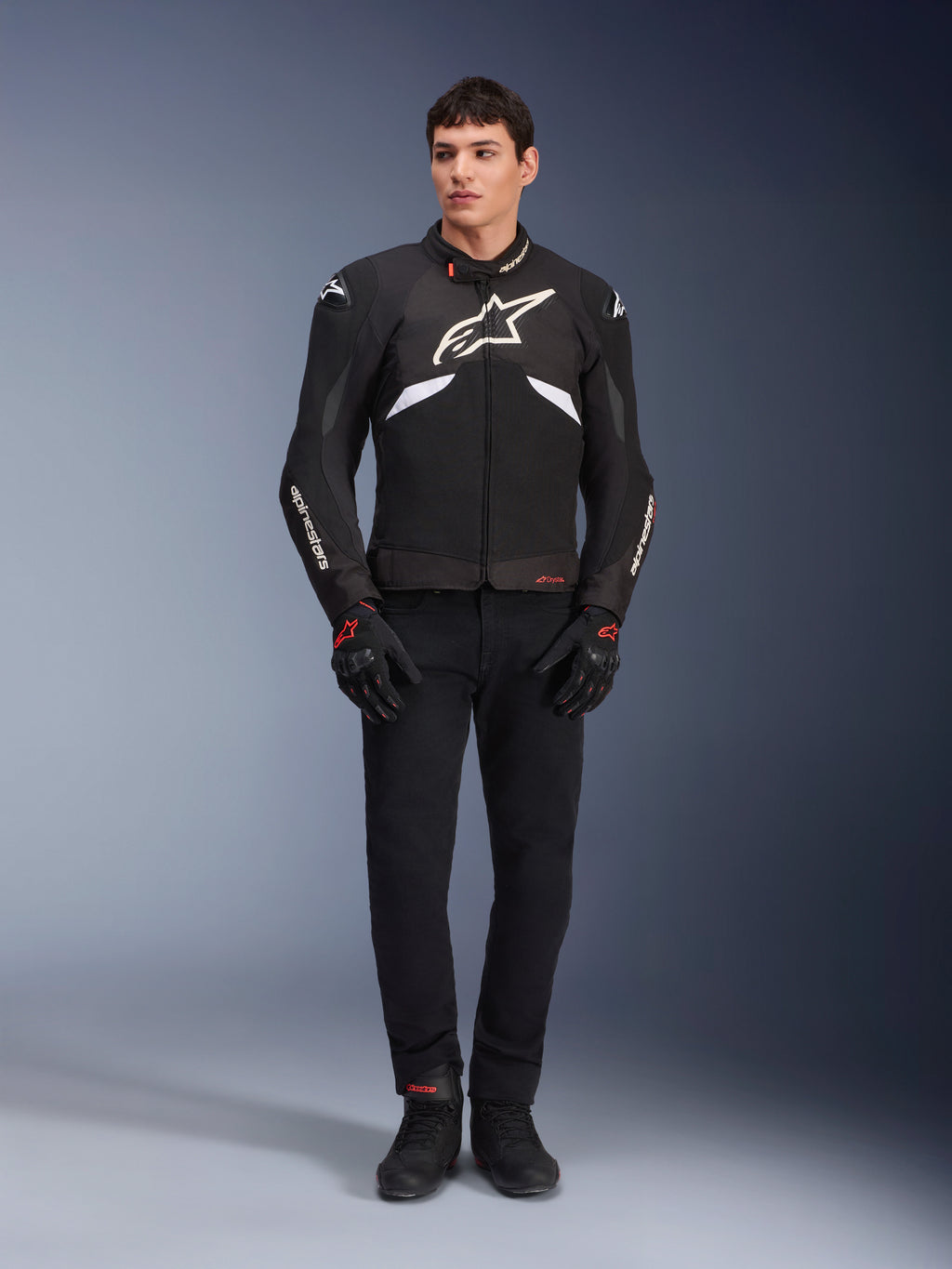 Männliches Model trägt die Alpinestars T-GP R V3 Drystar® Jacke in Schwarz und Weiß, wasserdichte Textil-Sportjacke mit großem weißem Alpinestars-Logo auf der Brust, kombiniert mit schwarzer Motorradhose und Handschuhen, professionelles Studioporträt, das die schlanke sportliche Silhouette hervorhebt.