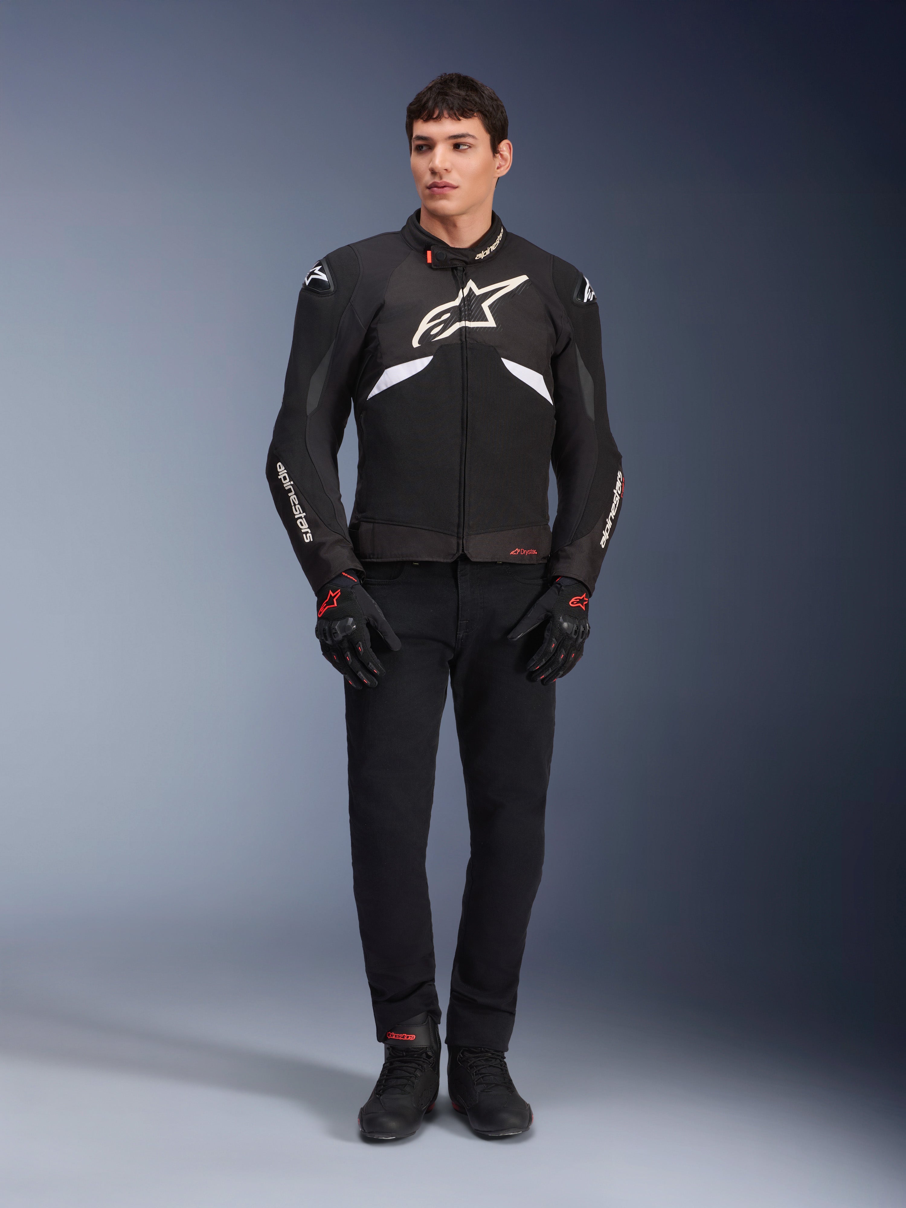 Männliches Model trägt die Alpinestars T-GP R V3 Drystar® Jacke in Schwarz und Weiß, wasserdichte Textil-Sportjacke mit großem weißem Alpinestars-Logo auf der Brust, kombiniert mit schwarzer Motorradhose und Handschuhen, professionelles Studioporträt, das die schlanke sportliche Silhouette hervorhebt.