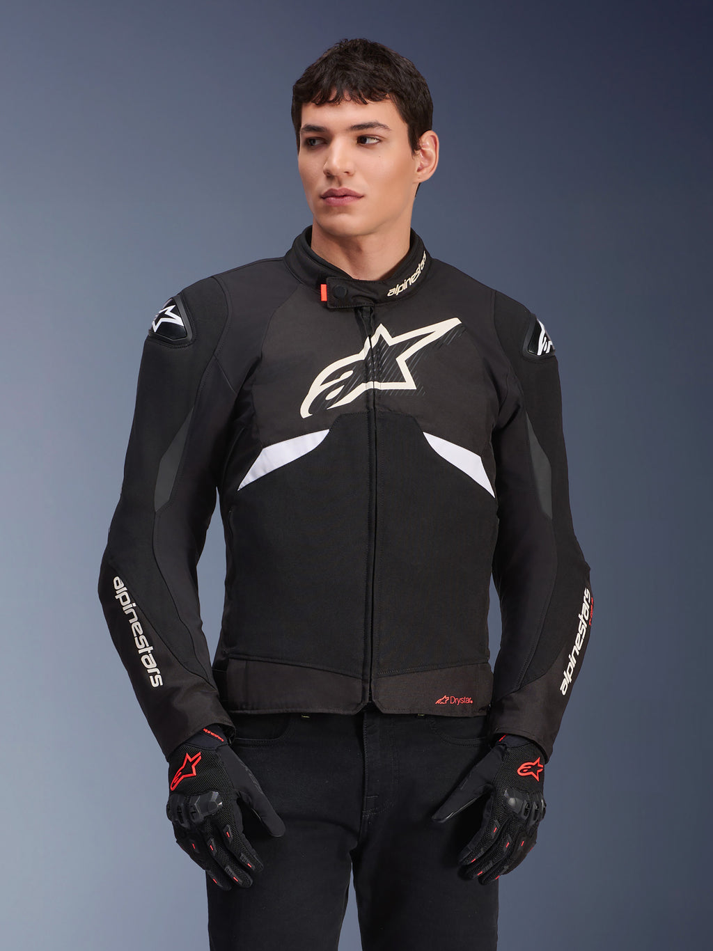 Alpinestars T-GP R V3 Drystar Jacke, wasserdichte Sport-Textiljacke, Schwarz und Weiß mit roten Akzenten, mit einem großen weißen Stern-Logo auf der Brust und weißem Branding auf den Ärmeln, getragen mit schwarz-roten Motorradhandschuhen