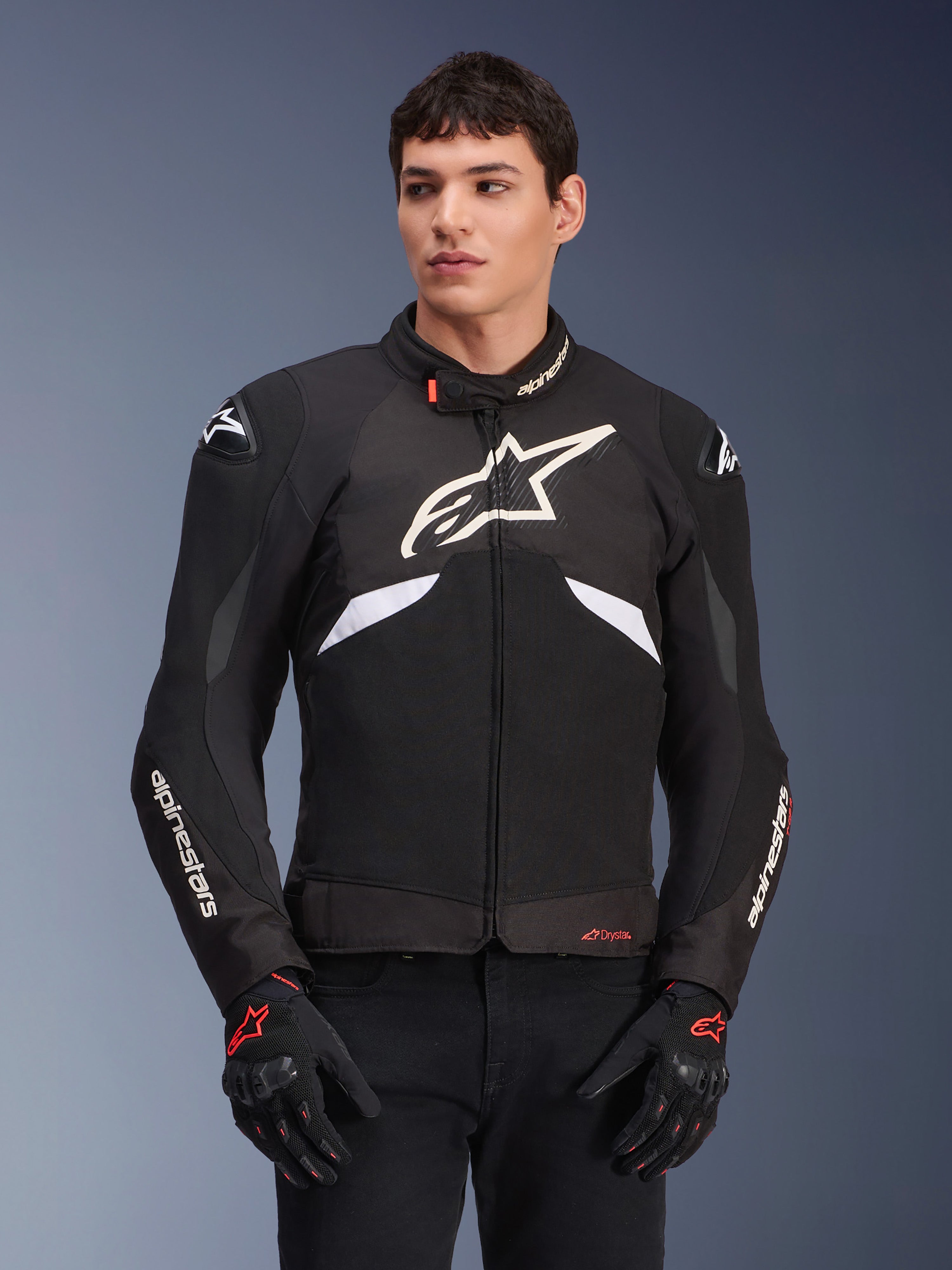 Alpinestars T-GP R V3 Drystar Jacke, wasserdichte Sport-Textiljacke, Schwarz und Weiß mit roten Akzenten, mit einem großen weißen Stern-Logo auf der Brust und weißem Branding auf den Ärmeln, getragen mit schwarz-roten Motorradhandschuhen