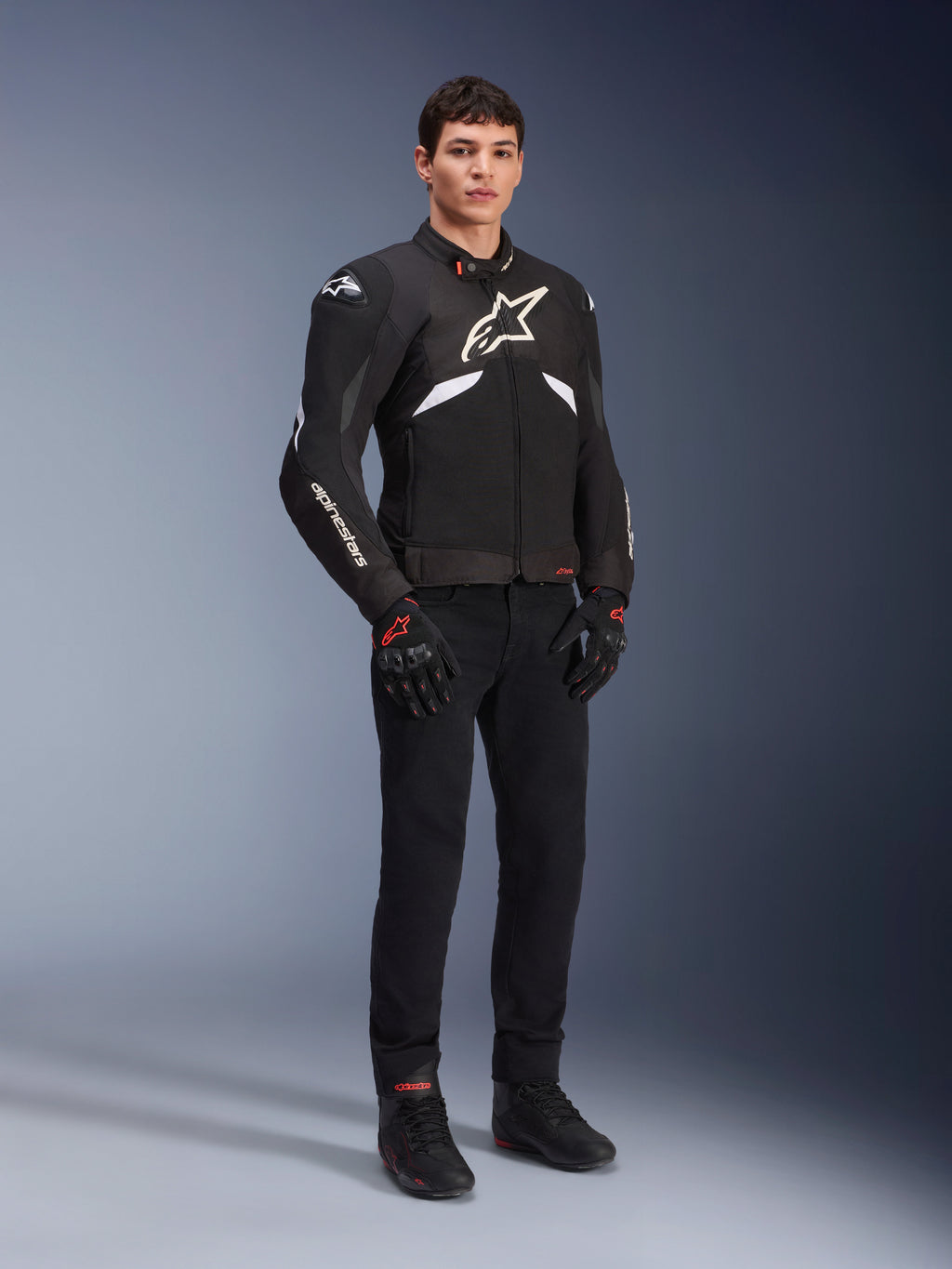 Alpinestars T-GP R V3 Drystar® Jacke in Schwarz und Weiß, getragen von einem Modell mit schwarzen Motorradhandschuhen und -hose, mit weißem Brustlogo, weißen Akzenten an den Ärmeln und schützenden Schulterschleifern, wasserdichte Textil-Sportjacke für die Straße