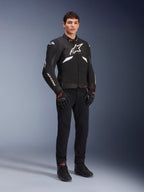 Alpinestars T-GP R V3 Drystar® Jacke in Schwarz und Weiß, getragen von einem Modell mit schwarzen Motorradhandschuhen und -hose, mit weißem Brustlogo, weißen Akzenten an den Ärmeln und schützenden Schulterschleifern, wasserdichte Textil-Sportjacke für die Straße
