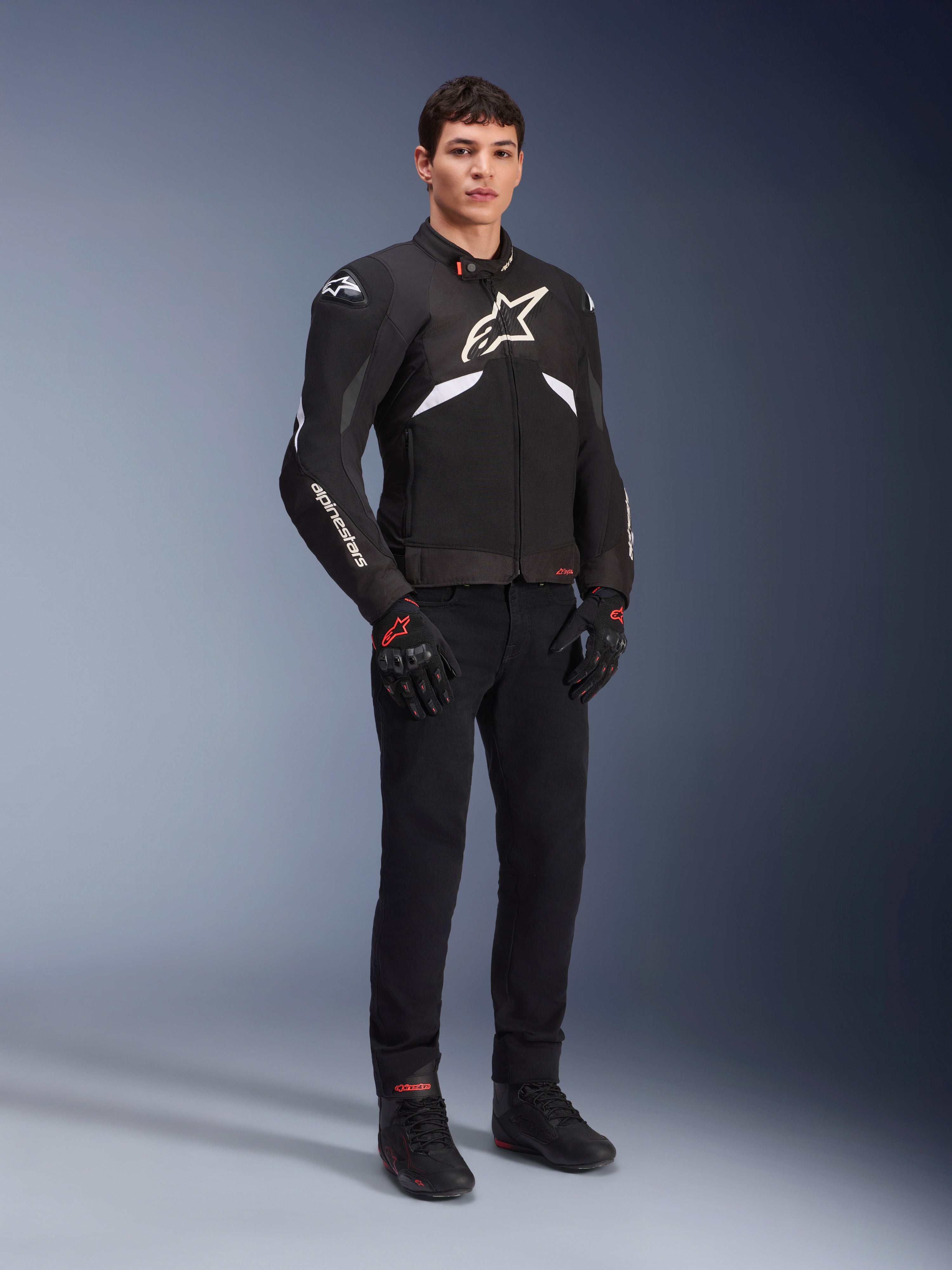 Alpinestars T-GP R V3 Drystar® Jacke in Schwarz und Weiß, getragen von einem Modell mit schwarzen Motorradhandschuhen und -hose, mit weißem Brustlogo, weißen Akzenten an den Ärmeln und schützenden Schulterschleifern, wasserdichte Textil-Sportjacke für die Straße