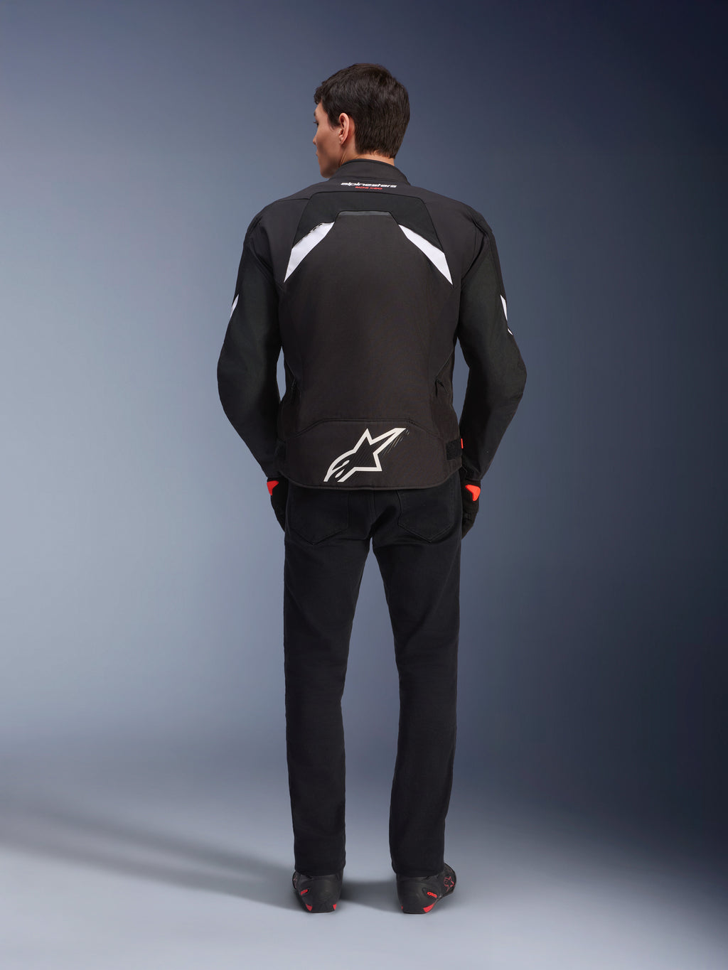 Alpinestars T-GP R V3 Drystar® Jacke, Schwarz und Weiß, Rückansicht, wasserdichte Textil-Motorradjacke mit einem großen weißen Stern-Logo auf dem unteren Rücken und weißen Akzenten an den Schultern, kombiniert mit schwarzer Hose und Stiefeln, rennsportinspiriertes sportliches Design