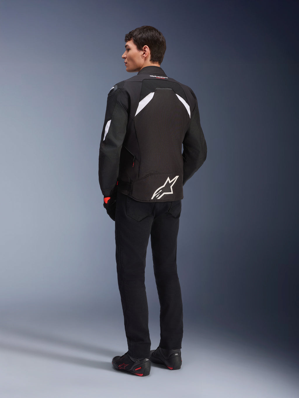 Männliches Model von hinten in der Alpinestars T-GP R V3 Drystar® Jacke in Schwarz und Weiß, mit weißen Schulterakzenten und einem großen weißen Logo am unteren Rücken, kombiniert mit schwarzer Motorradhose und Stiefeln, sportliche Textil-Motorradjacke.