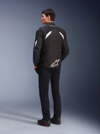Männliches Model von hinten in der Alpinestars T-GP R V3 Drystar® Jacke in Schwarz und Weiß, mit weißen Schulterakzenten und einem großen weißen Logo am unteren Rücken, kombiniert mit schwarzer Motorradhose und Stiefeln, sportliche Textil-Motorradjacke.