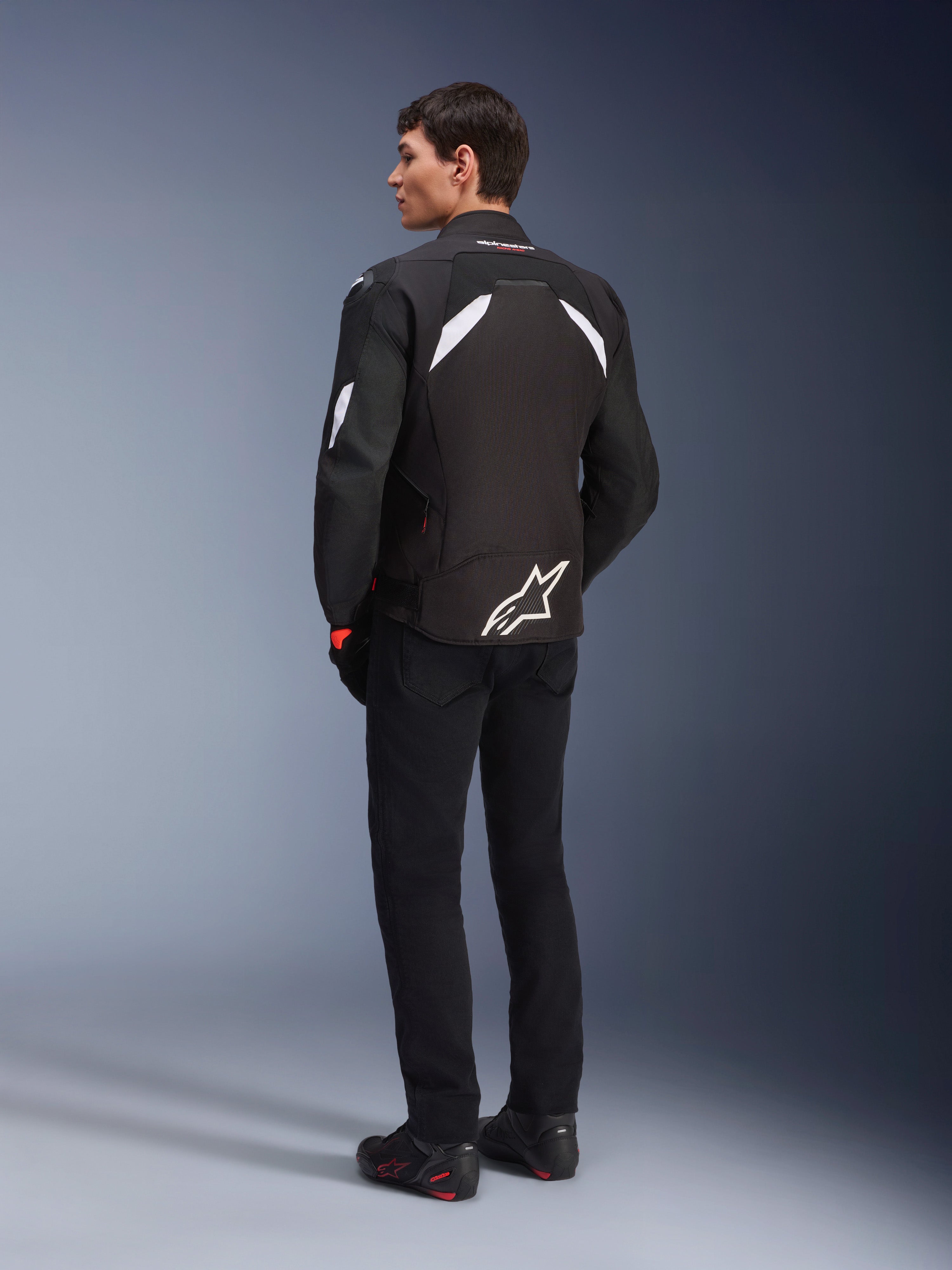 Männliches Model von hinten in der Alpinestars T-GP R V3 Drystar® Jacke in Schwarz und Weiß, mit weißen Schulterakzenten und einem großen weißen Logo am unteren Rücken, kombiniert mit schwarzer Motorradhose und Stiefeln, sportliche Textil-Motorradjacke.