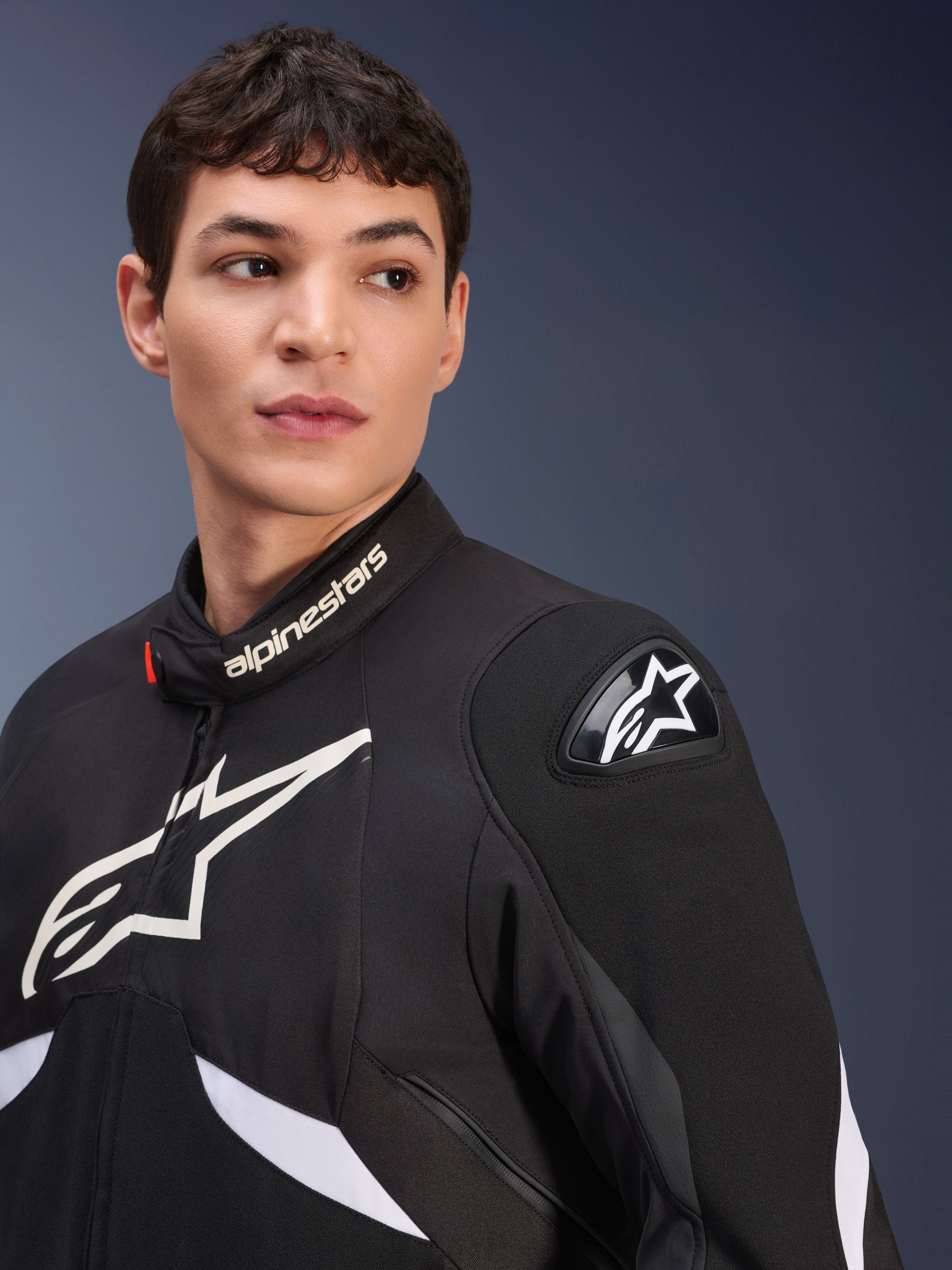 Alpinestars T-GP R V3 Drystar® Jacke, Schwarz und Weiß, getragen von einem männlichen Model, mit weißen Alpinestars Logos an Kragen und Brust, schwarzer DFS Schulterschleifer mit weißem Stern, detaillierte Lifestyle-Aufnahme einer wasserdichten Sport-Textiljacke