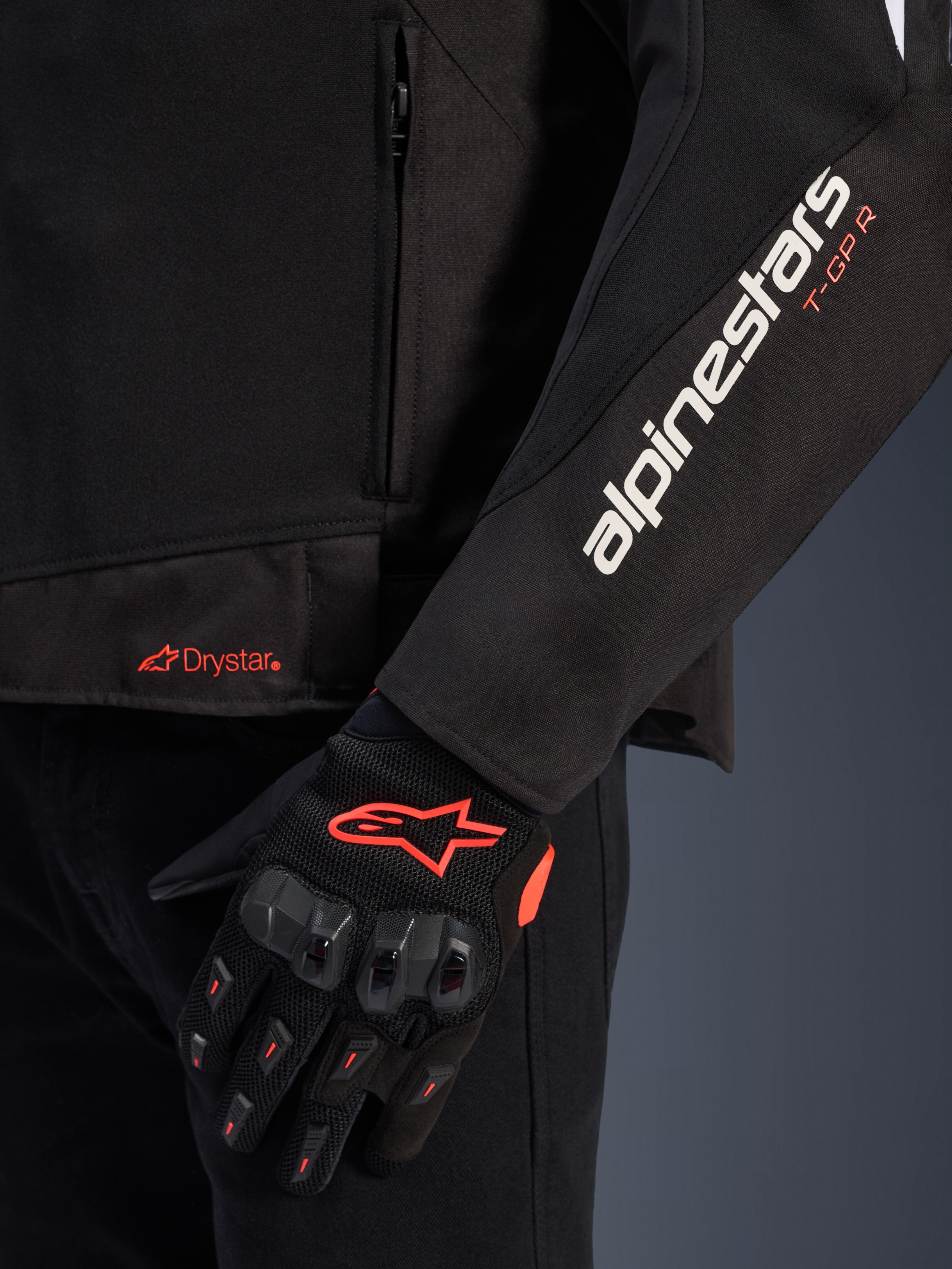 Alpinestars T-GP R V3 Drystar® Jacke, schwarze Textil-Motorradjacke, Nahaufnahme mit weißem Alpinestars- und orangefarbenem T-GP R-Branding am Ärmel, orangefarbenes Drystar-Logo am Saum, kombiniert mit schwarzen Mesh-Schutzhandschuhen mit roten Akzenten.