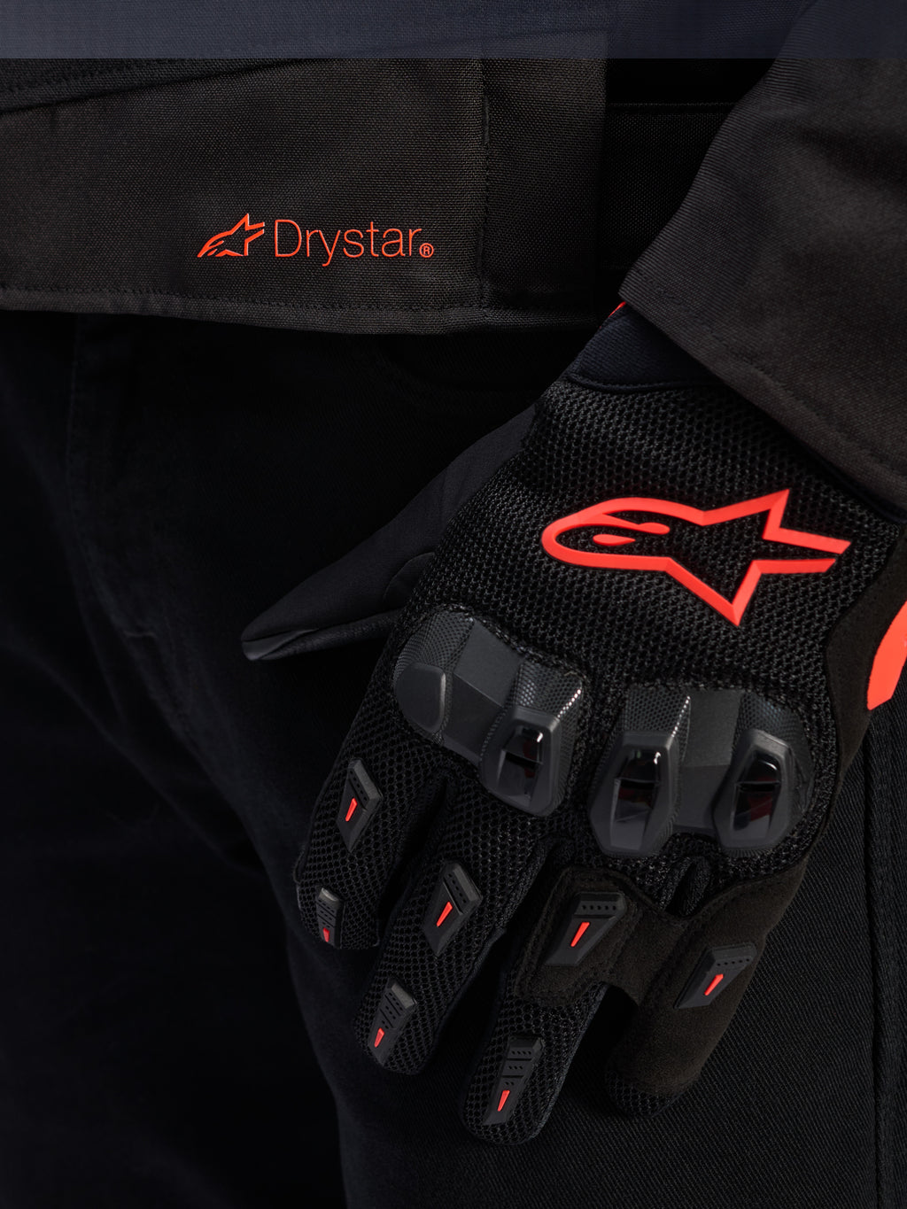 Nahaufnahme des Taillenbereichs der Alpinestars T-GP R V3 Drystar® Jacke mit rotem Drystar-Logo, gezeigt mit einem schwarzen Motorradhandschuh mit rotem Alpinestars Stern-Logo, Mesh-Gewebe und verstärktem Knöchelschutz.