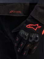 Nahaufnahme des Taillenbereichs der Alpinestars T-GP R V3 Drystar® Jacke mit rotem Drystar-Logo, gezeigt mit einem schwarzen Motorradhandschuh mit rotem Alpinestars Stern-Logo, Mesh-Gewebe und verstärktem Knöchelschutz.