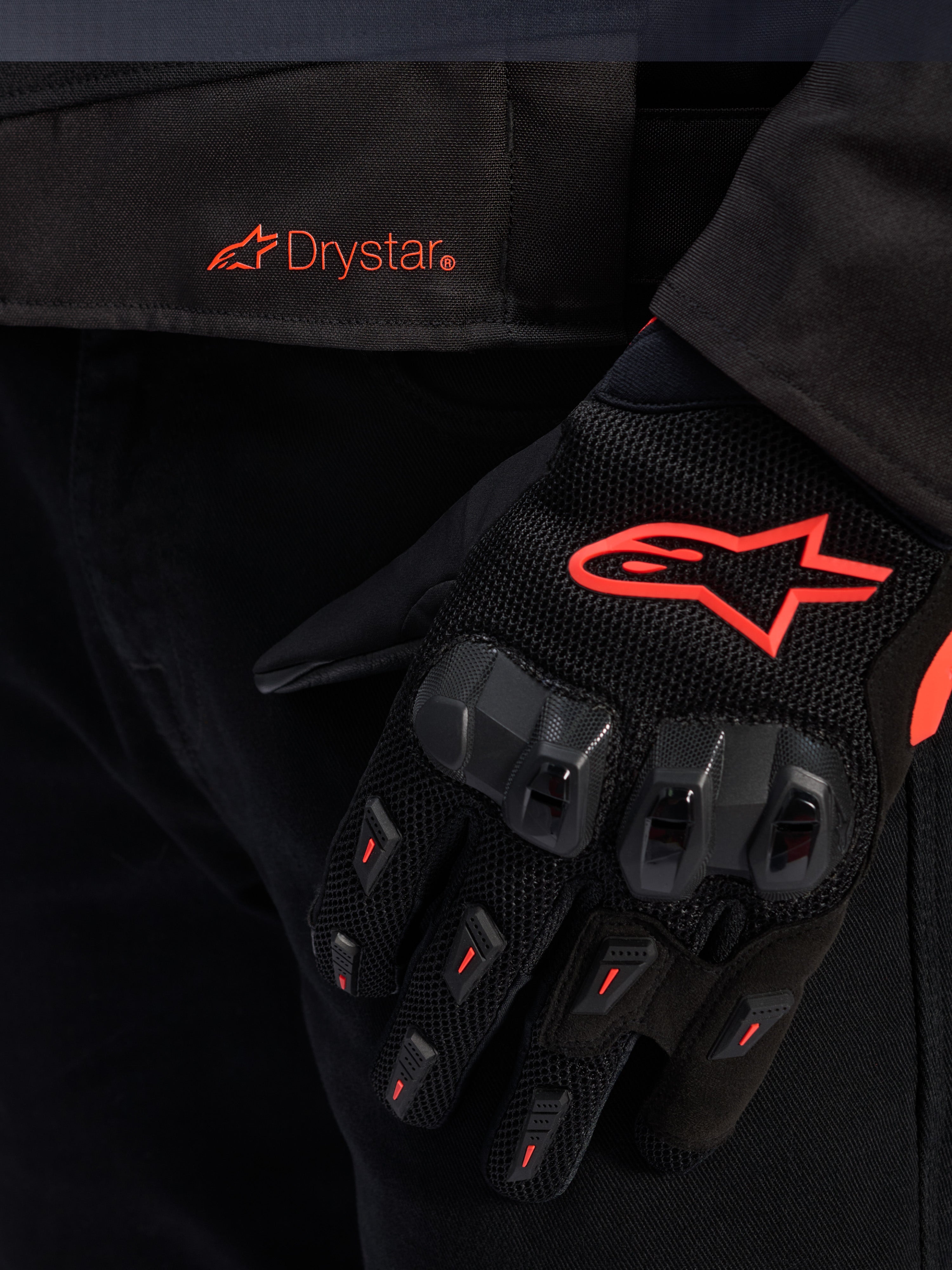 Nahaufnahme des Taillenbereichs der Alpinestars T-GP R V3 Drystar® Jacke mit rotem Drystar-Logo, gezeigt mit einem schwarzen Motorradhandschuh mit rotem Alpinestars Stern-Logo, Mesh-Gewebe und verstärktem Knöchelschutz.