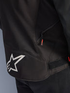 Alpinestars T-GP R V3 Drystar® Jacke, Schwarz-Weiß, Detailansicht des unteren Rückenbereichs mit einem großen weißen Alpinestars Stern-Logo, einer seitlichen Reißverschlusstasche mit roter Zuglasche und einem Klettverschluss-Riemen zur Taillenverstellung, strapazierfähiges Textilmaterial.