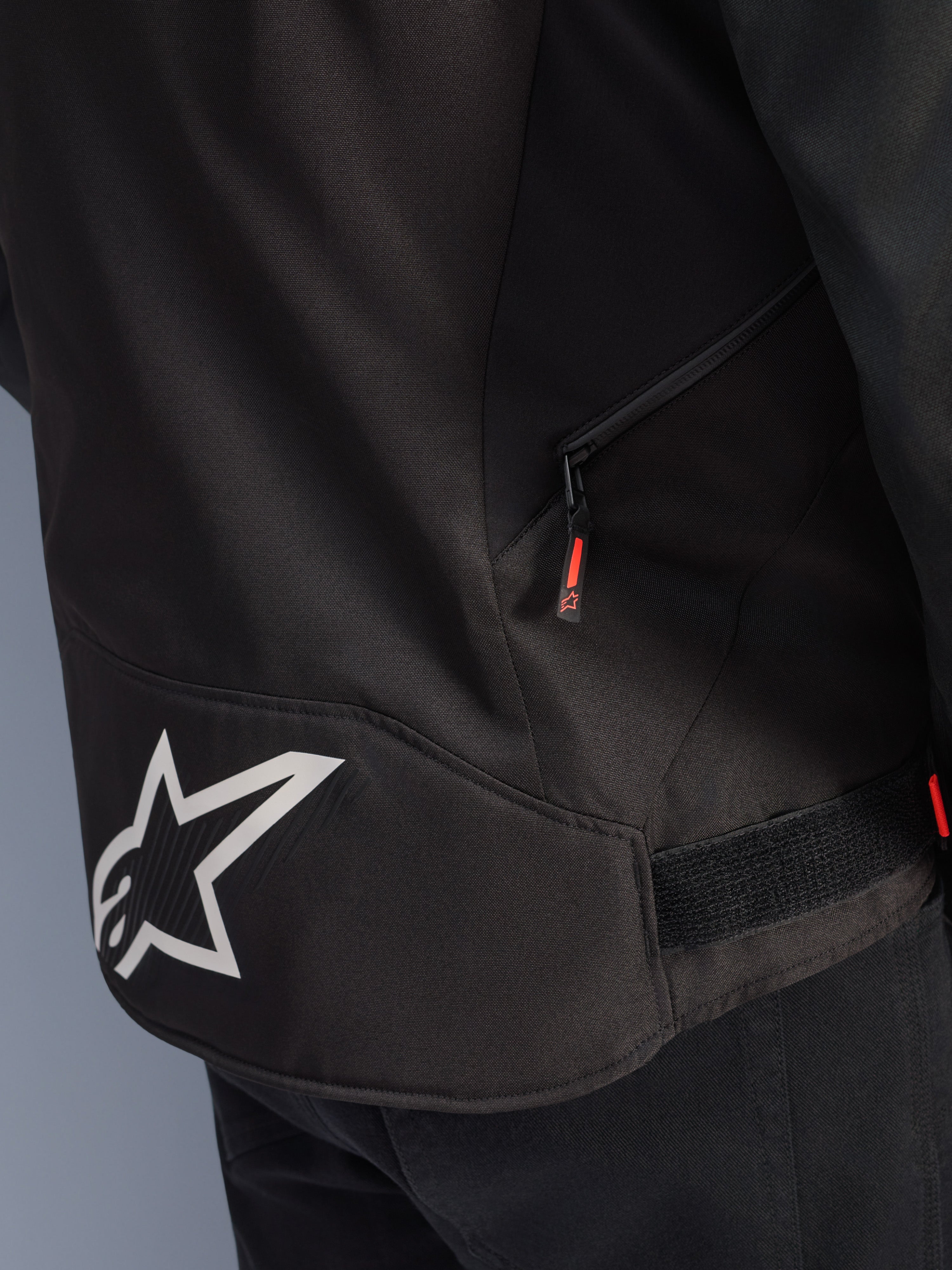 Alpinestars T-GP R V3 Drystar® Jacke, Schwarz-Weiß, Detailansicht des unteren Rückenbereichs mit einem großen weißen Alpinestars Stern-Logo, einer seitlichen Reißverschlusstasche mit roter Zuglasche und einem Klettverschluss-Riemen zur Taillenverstellung, strapazierfähiges Textilmaterial.