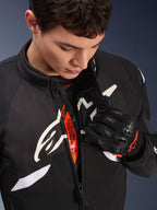 Alpinestars T-GP R V3 Drystar® Jacke, schwarz-weiße Motorrad-Sporttextiljacke, teilweise geöffnet mit sichtbarem orangefarbenem „Alpinestars Protects“-Innenband, getragen mit schwarz-roten Mesh-Handschuhen, vom Rennsport abgeleitete Schutzausrüstung.