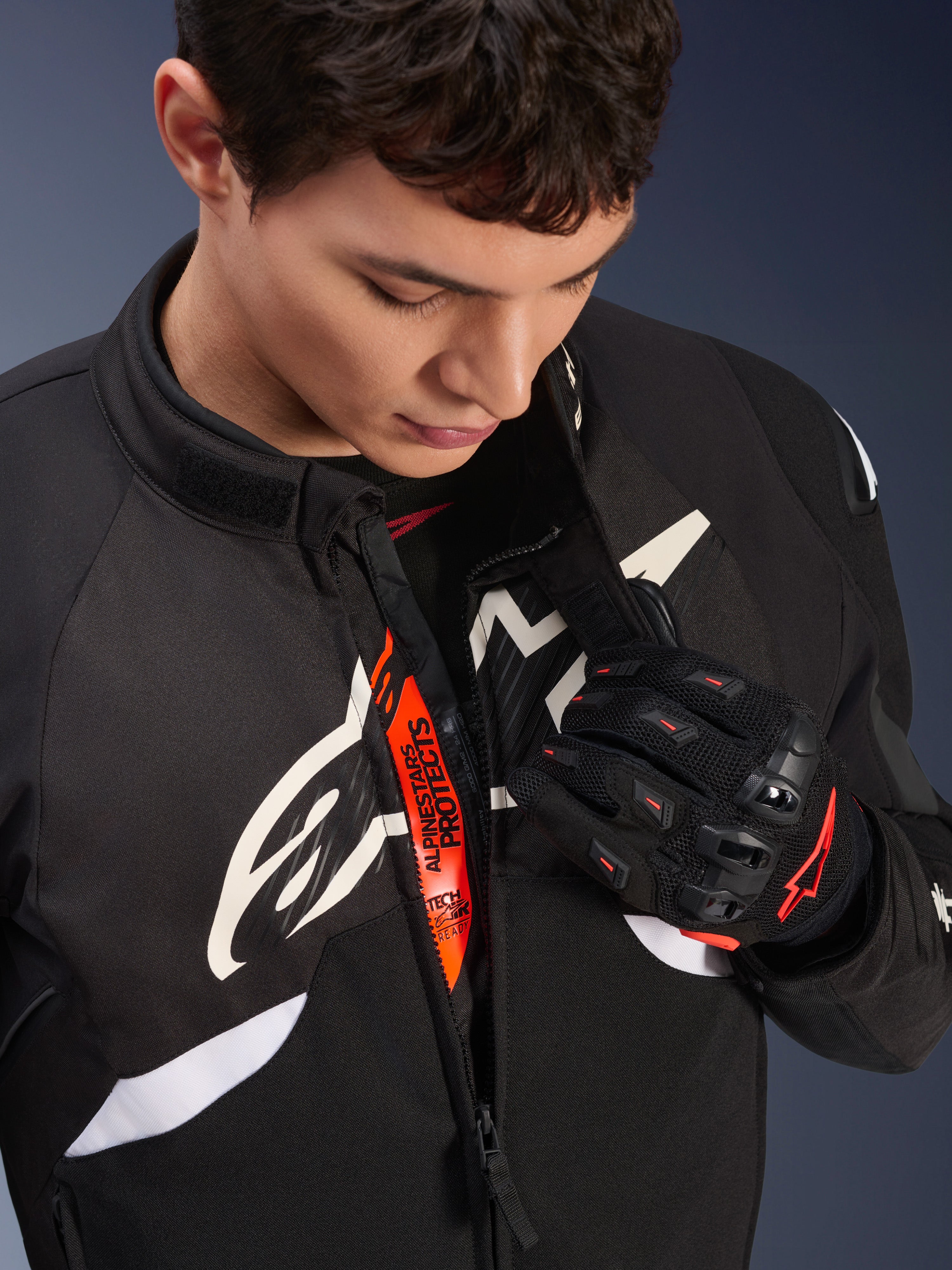 Alpinestars T-GP R V3 Drystar® Jacke, schwarz-weiße Motorrad-Sporttextiljacke, teilweise geöffnet mit sichtbarem orangefarbenem „Alpinestars Protects“-Innenband, getragen mit schwarz-roten Mesh-Handschuhen, vom Rennsport abgeleitete Schutzausrüstung.