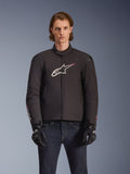 Alpinestars SMX Wasserdichte Jacke, schwarze Textil-Motorradjacke mit weißem und rotem Stern-Logo auf der Brust, getragen von einem Model mit schwarzen Handschuhen und blauen Jeans, mit externen Schulterschleifern und sportlicher Passform.