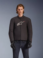 Alpinestars SMX Wasserdichte Jacke, schwarze Textil-Motorradjacke mit weißem und rotem Stern-Logo auf der Brust, getragen von einem Model mit schwarzen Handschuhen und blauen Jeans, mit externen Schulterschleifern und sportlicher Passform.