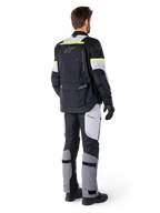 Rückansicht eines Fahrers in der Alpinestars Bogotá Pro Drystar® Jacke in Black Ice Gray Yellow Fluo, kombiniert mit passender Adventure-Tourenhose, mit einer großen Rückentasche und neongelben Akzenten, Ganzjahres-Textil-Motorradausrüstung.