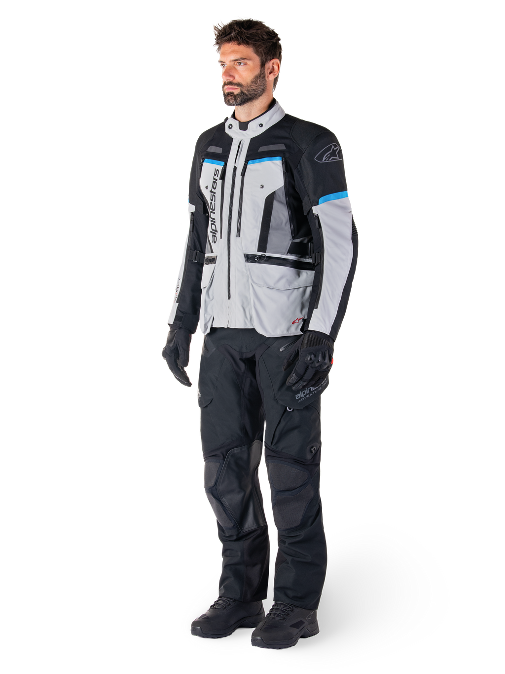 Männlicher Motorradfahrer in der Alpinestars Bogotá Pro Drystar® Jacke in Eisgrau, Schwarz und Cyan, kombiniert mit schwarzer technischer Motorradhose und Handschuhen, Adventure-Touring-Ausrüstung für Performance bei jedem Wetter.