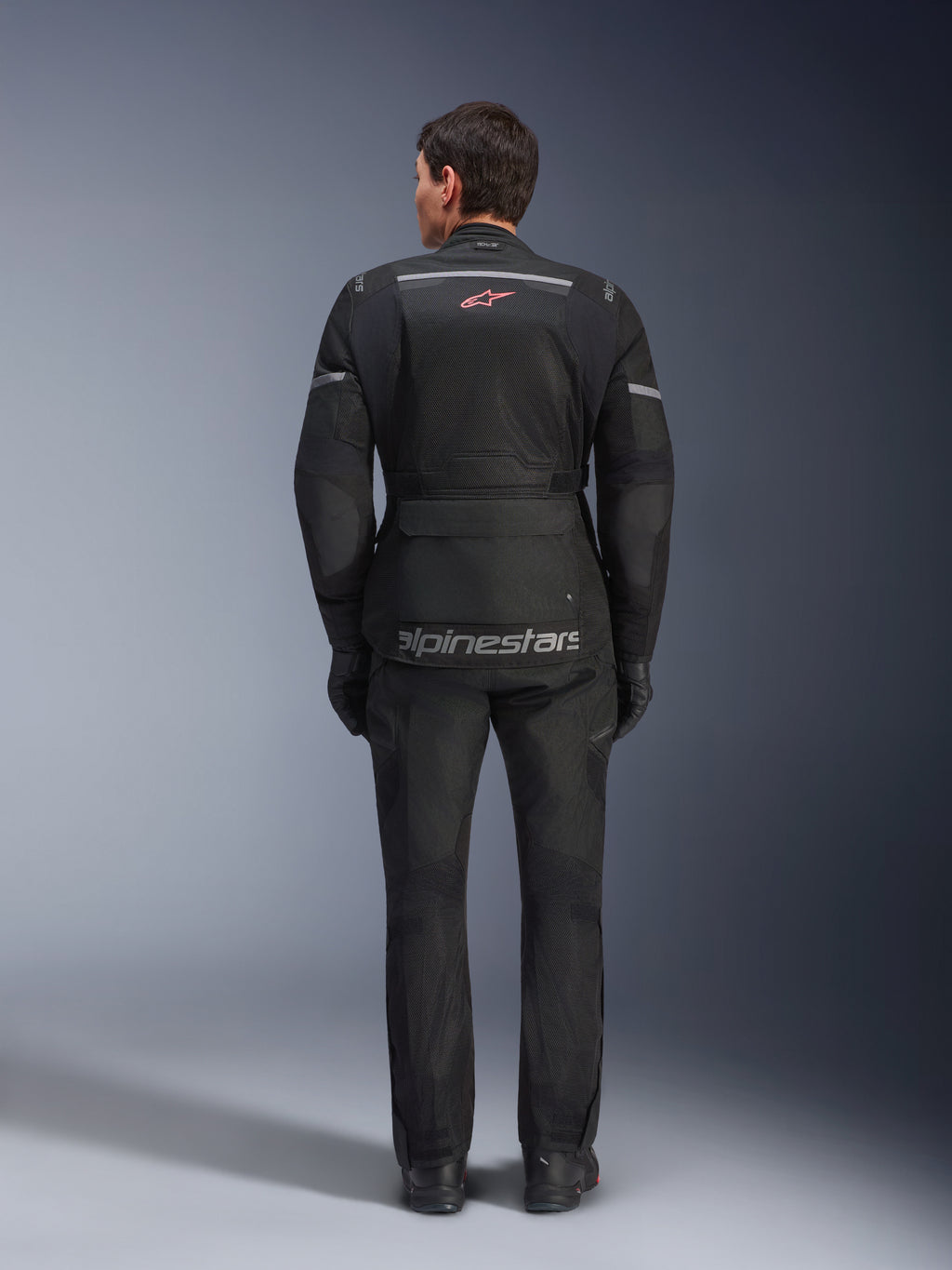 Alpinestars Andes Air Drystar® Jacke und Hose, schwarzes Motorrad-Tourenoutfit, Rückansicht des Fahrers, mit Mesh-Einsätzen, rotem Stern-Logo und reflektierendem Alpinestars-Branding auf der Jacke, getragen mit schwarzen Handschuhen und Stiefeln.