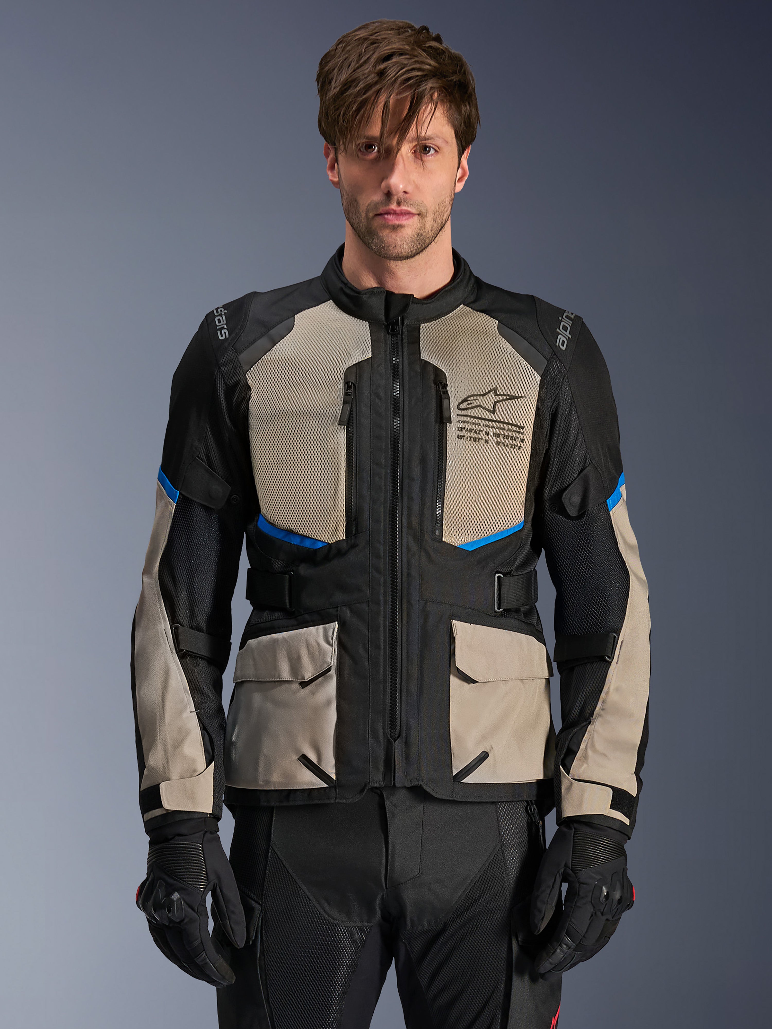 Alpinestars Andes Air Drystar Jacke, Adventure-Touringjacke, Schwarz, Laurel Gray und Bright Blue, getragen von einem Modell mit schwarzen Handschuhen, mit großen Mesh-Einsätzen im Brustbereich für Luftzirkulation, vorderen Cargotaschen und blauen Akzenten, schützende Motorrad-Tourenausrüstung