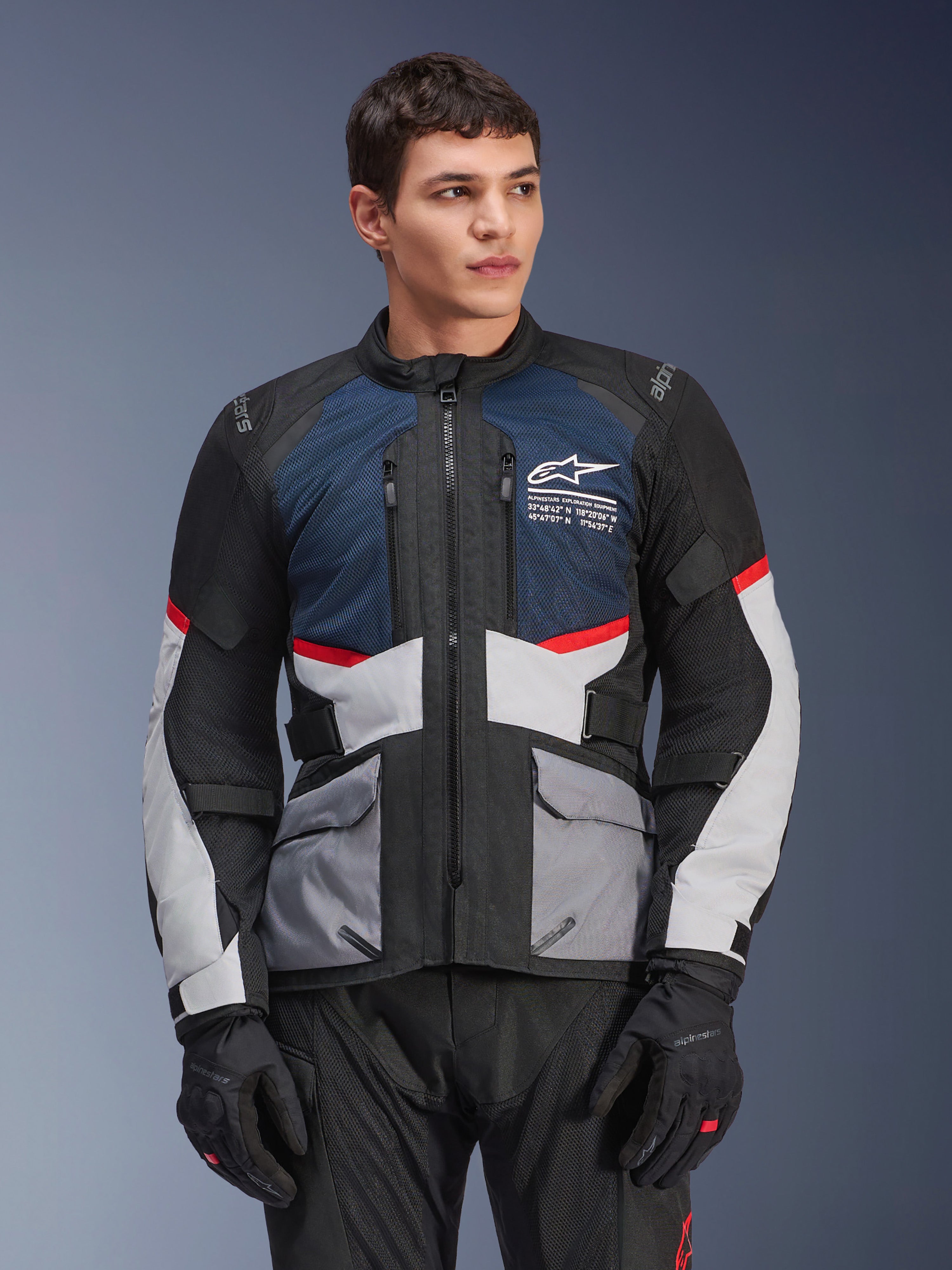 Alpinestars Andes Air Drystar® Jacke, Motorrad-Tourenjacke, Dunkelblau, Schwarz und Eisgrau mit roten Akzenten, getragen von einem männlichen Model mit schwarzen Handschuhen, verfügt über Mesh-Belüftungseinsätze und praktische Vordertaschen, moderne Abenteuer- und Straßenausrüstung