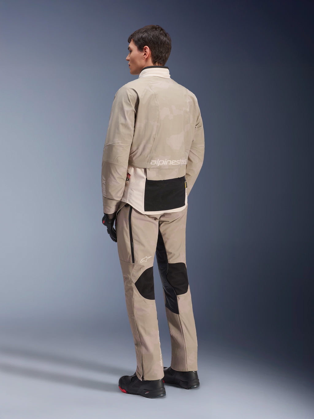 Model from the rear wearing the AMT-10LAB Drystar® XF Jacket and matching pants, White Sand Aluminum Camo, adventure motorcycle gear, featuring black utility panels and Alpinestars branding, worn with black riding boots and gloves, coordinated outfit for all-weather touring