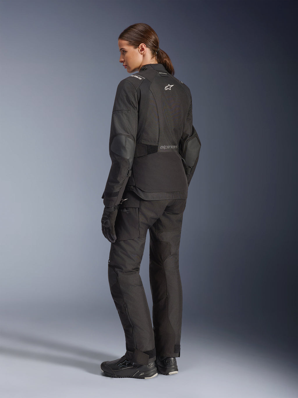 Motorradfahrerin in der Alpinestars Stella Andes V4 Drystar® Jacke, schwarze Adventure-Motorradjacke, Dreiviertel-Rückansicht, kombiniert mit passender Textilhose und Handschuhen, weiße Alpinestars-Logos am Rücken und an der Taille, verstärkte Schutzpaneele, verlängerter Saum am Rücken.