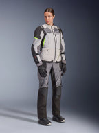 Frau trägt Alpinestars Stella Andes V4 Drystar® Jacke und passende Hose, Ice Gray und Dark Gray mit neongelben Akzenten, komplettes Adventure-Touring-Bekleidungsset mit schwarzen Handschuhen und Stiefeln, Ganzkörper-Studioaufnahme.