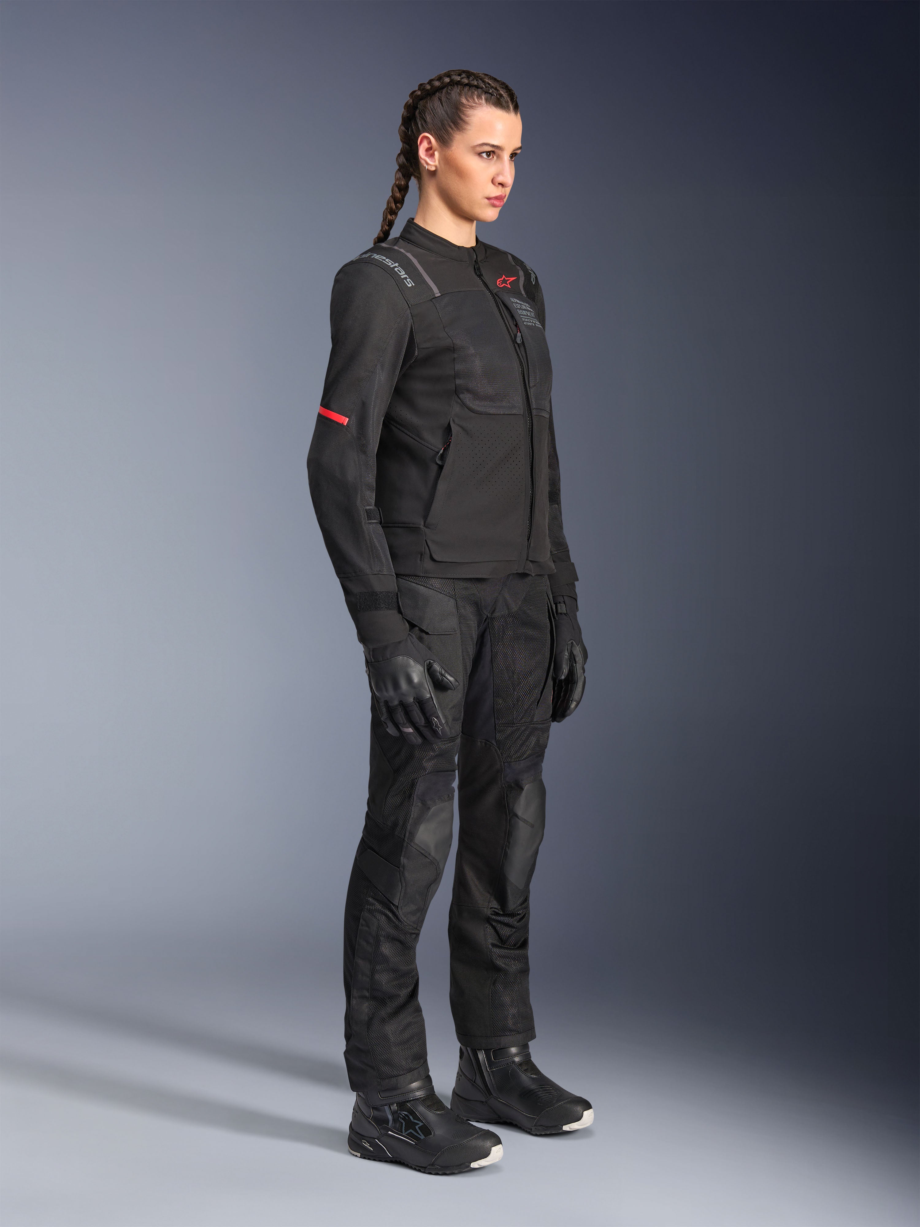 Weibliches Model trägt die Alpinestars Stella ST-2 Air Jacke in Schwarz, kombiniert mit passender schwarzer Motorradhose, Handschuhen und Stiefeln, mit Mesh-Belüftungseinsätzen und dezenten roten Details, elegantes modernes Touren-Outfit